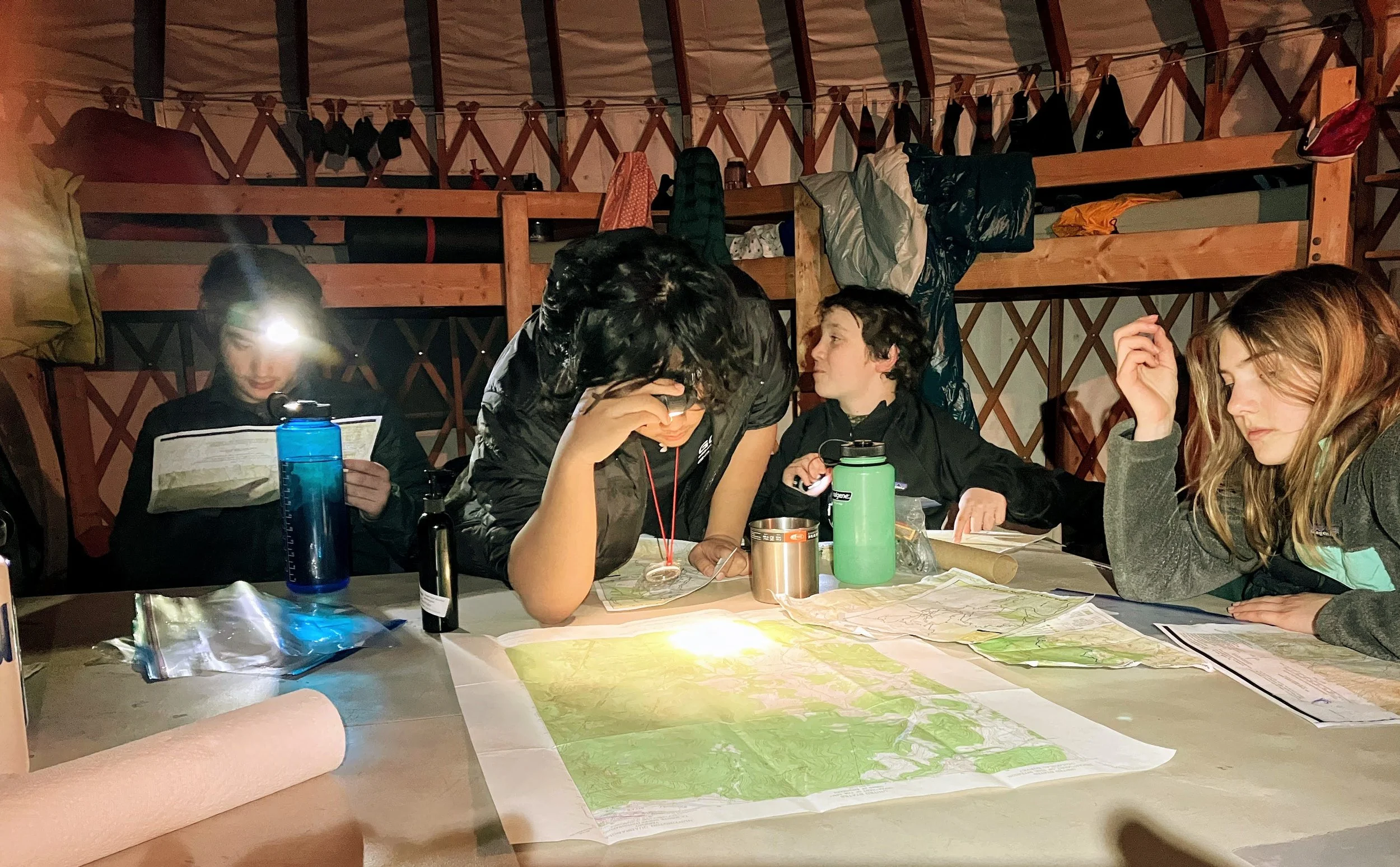 Group of five people sitting at a table with maps, in a yurt, during a camping or outdoor trip, with backpacks and gear around.