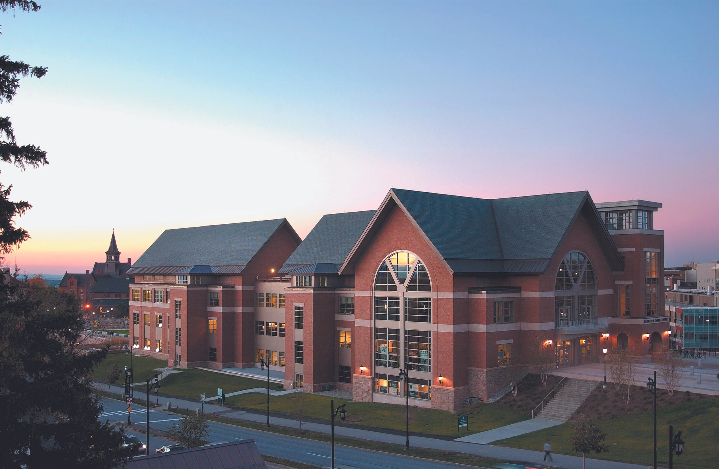 A large brick building with multiple stories and arched windows, situated near a street with lamp posts and sidewalks, during sunset with a pastel-colored sky.