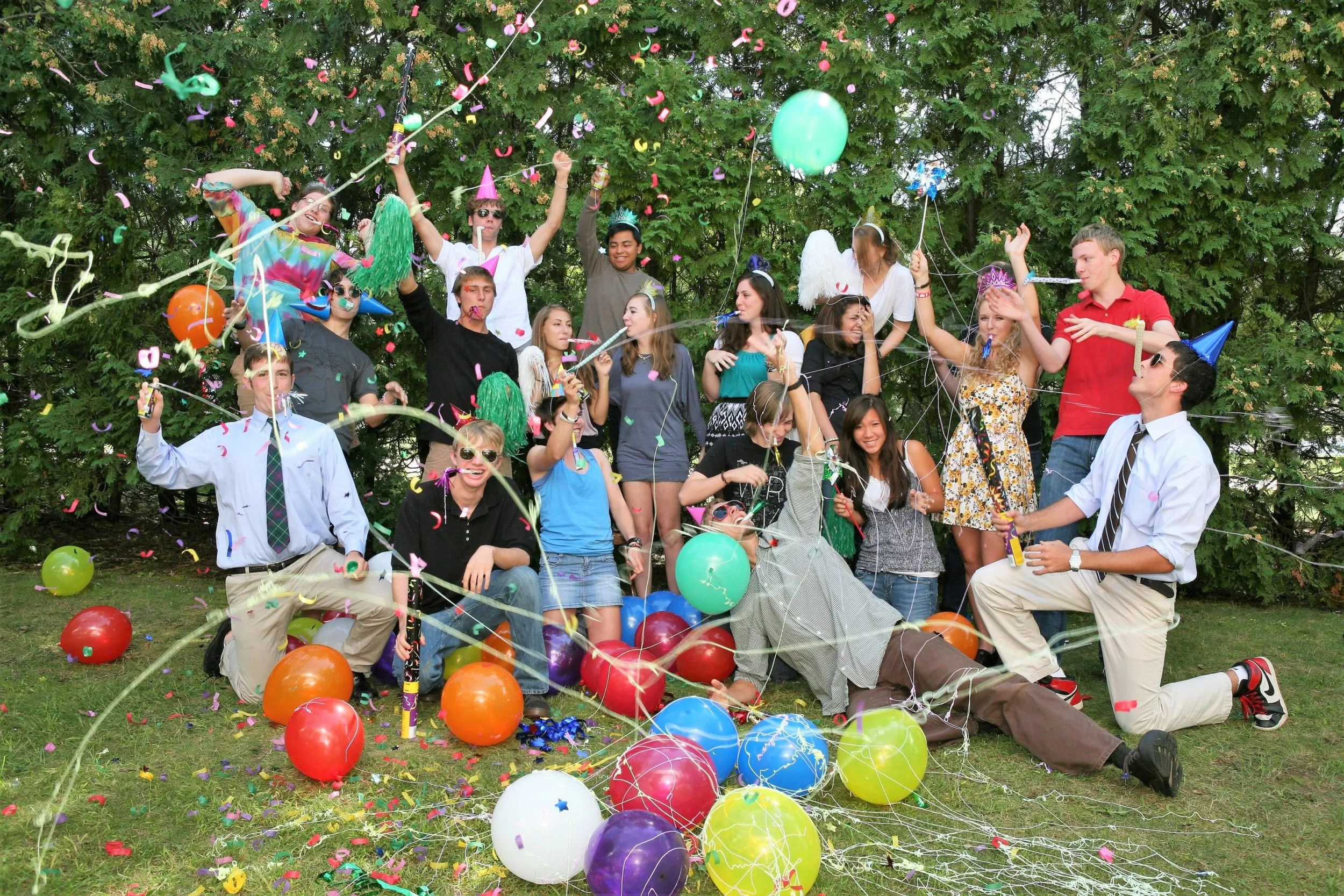 A group of young people celebrating outdoors with balloons, confetti, and party hats, enjoying a festive gathering.