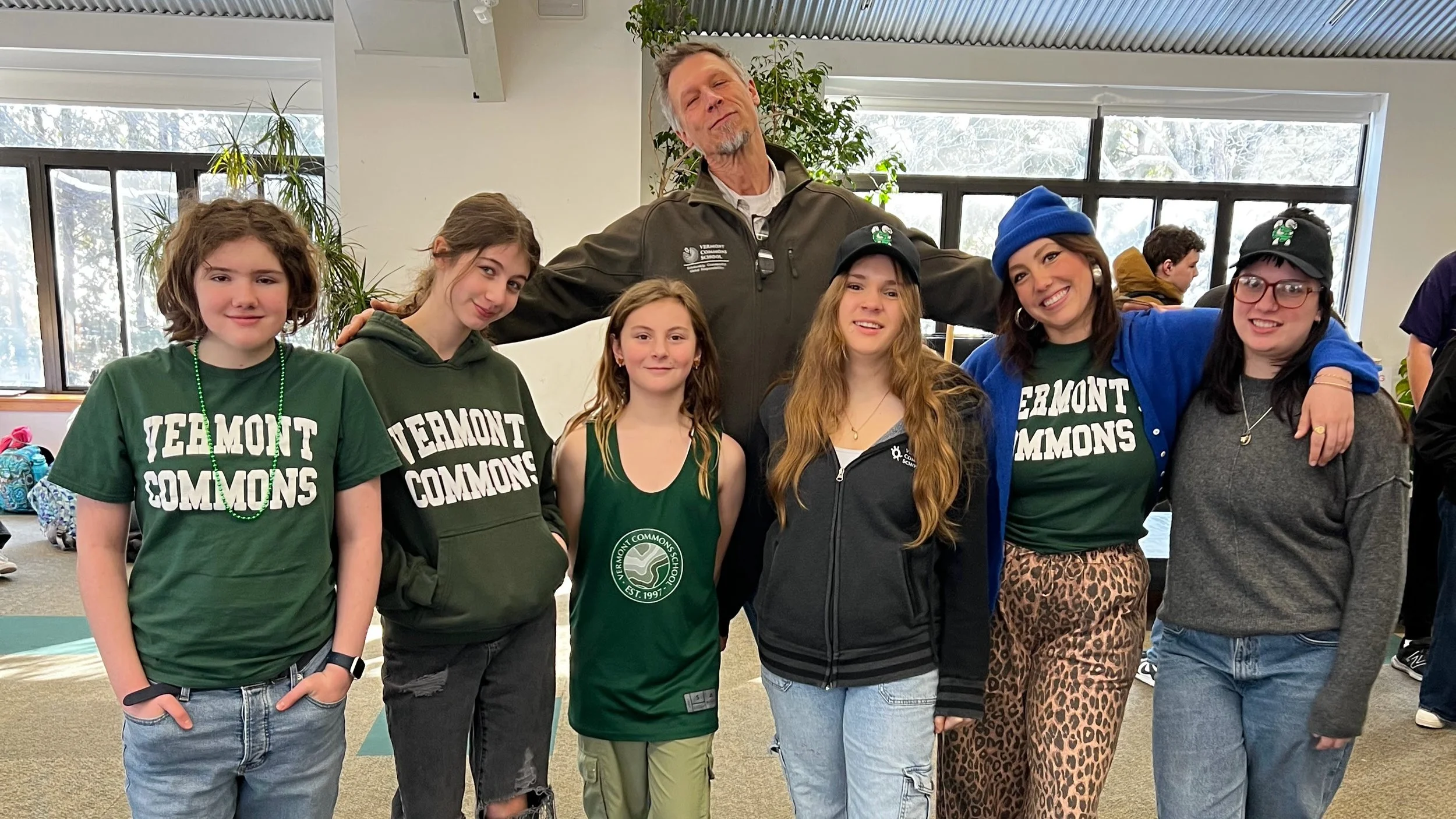Group of seven people, five young women and one older man, standing indoors with large windows in the background. The young women are wearing green shirts with