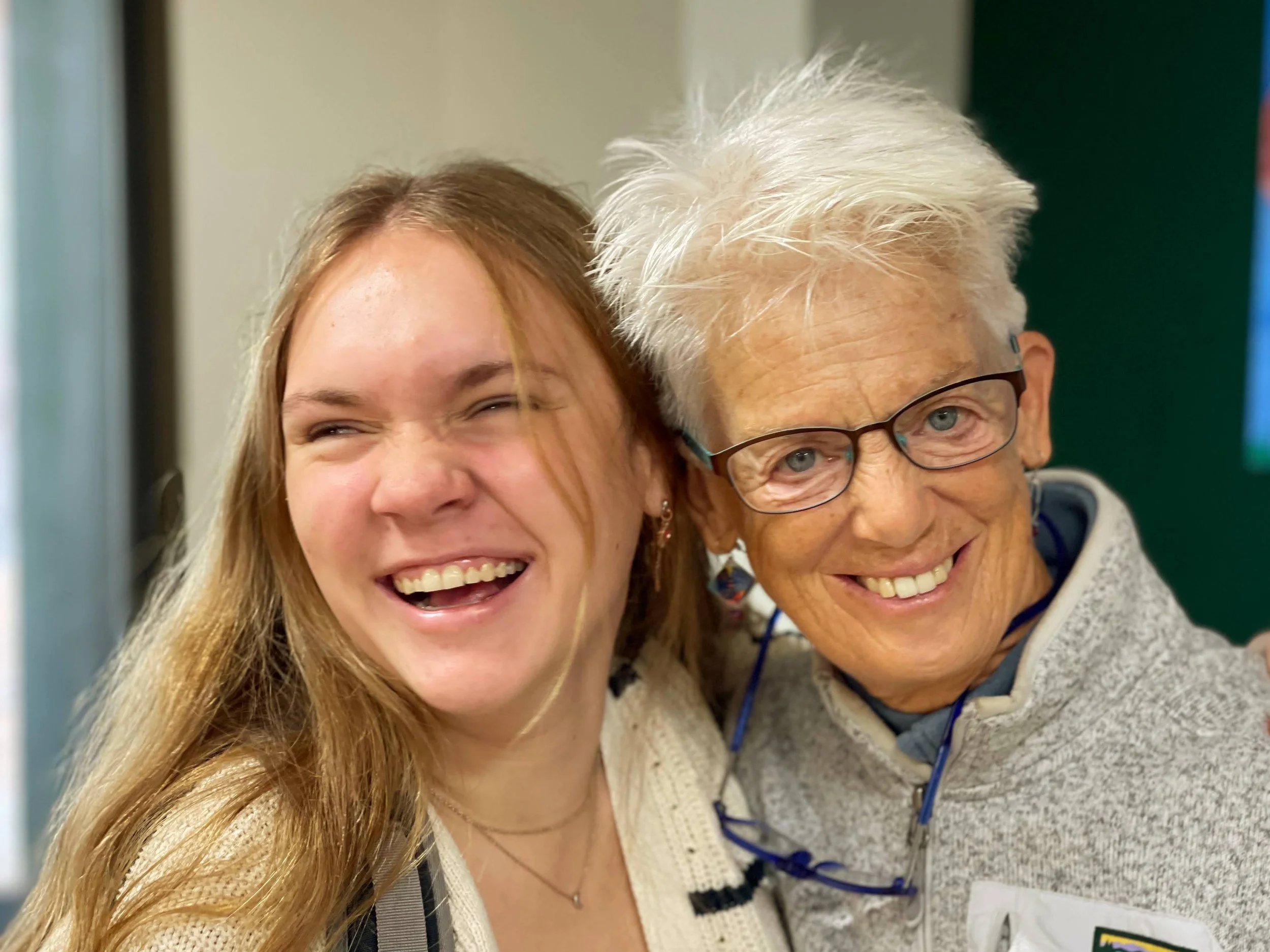 Two women smiling and close together, one with long red hair and the other with short white hair and glasses.