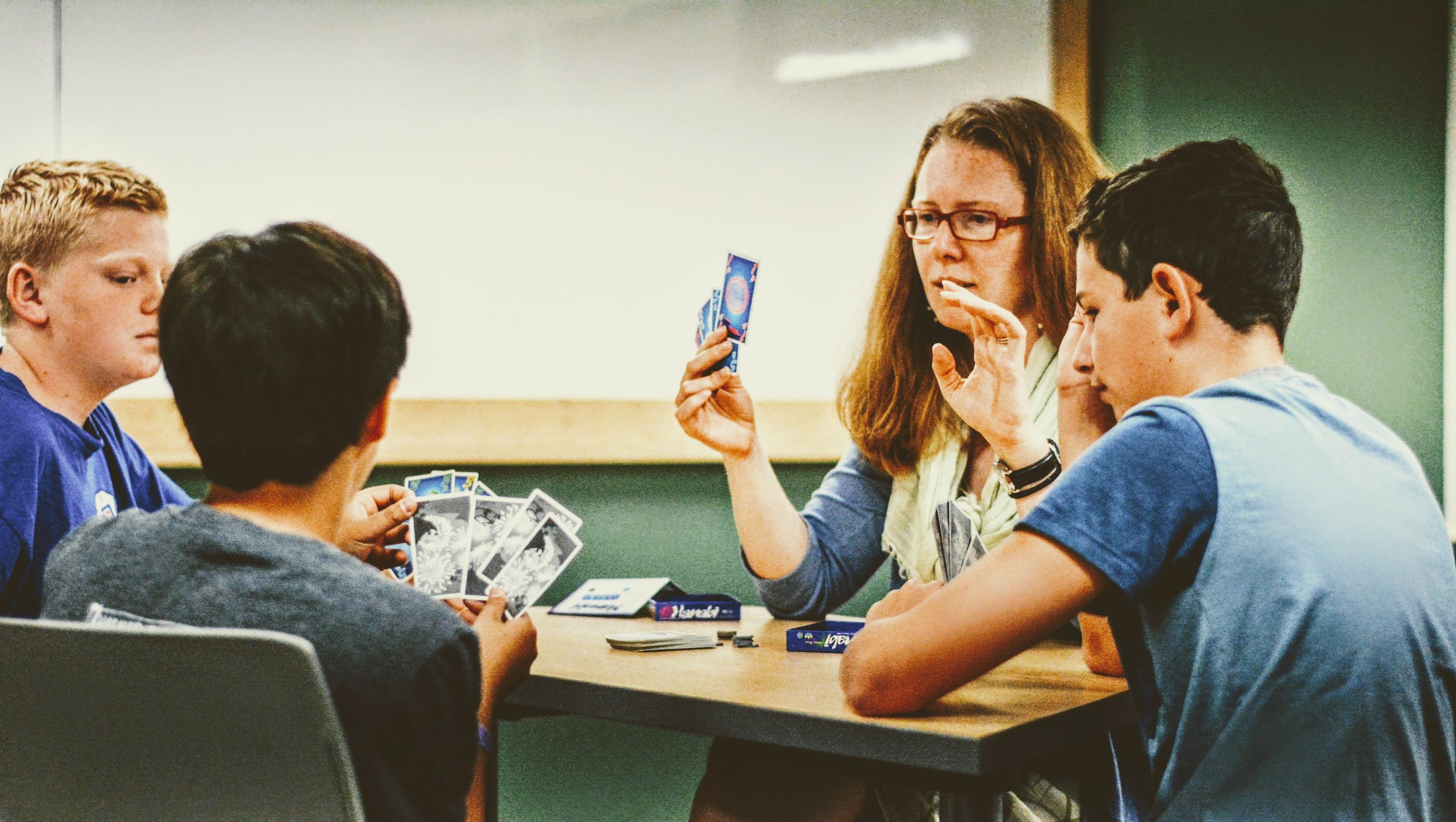 Four children and a woman are playing a card game at a table in a classroom, with a whiteboard in the background.