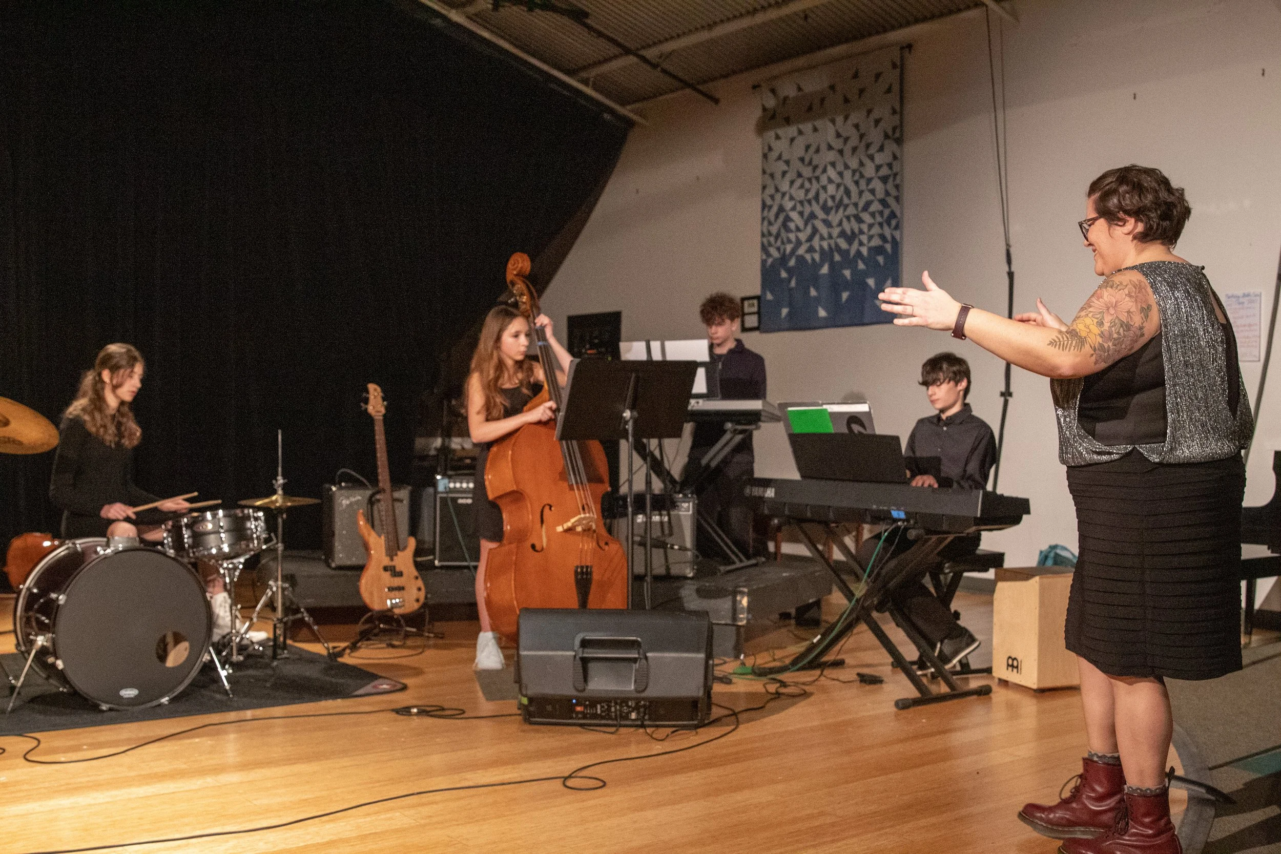 A music teacher instructs a band of young musicians during a rehearsal or performance, with a drum set, bass guitar, double bass, keyboard, and other equipment on stage.