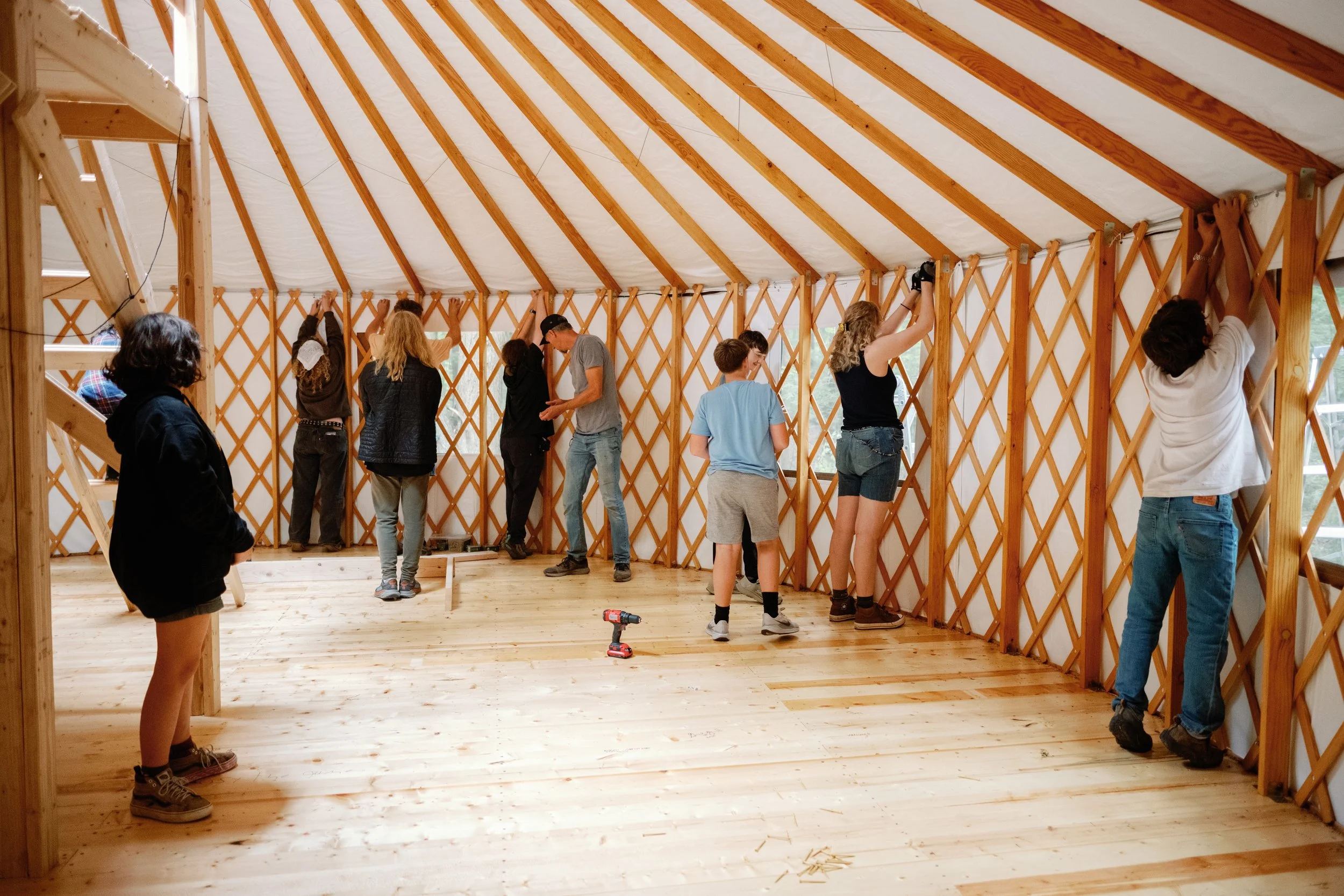People working together to build the wooden wall of a yurt, assembling lattice walls inside a wooden frame.
