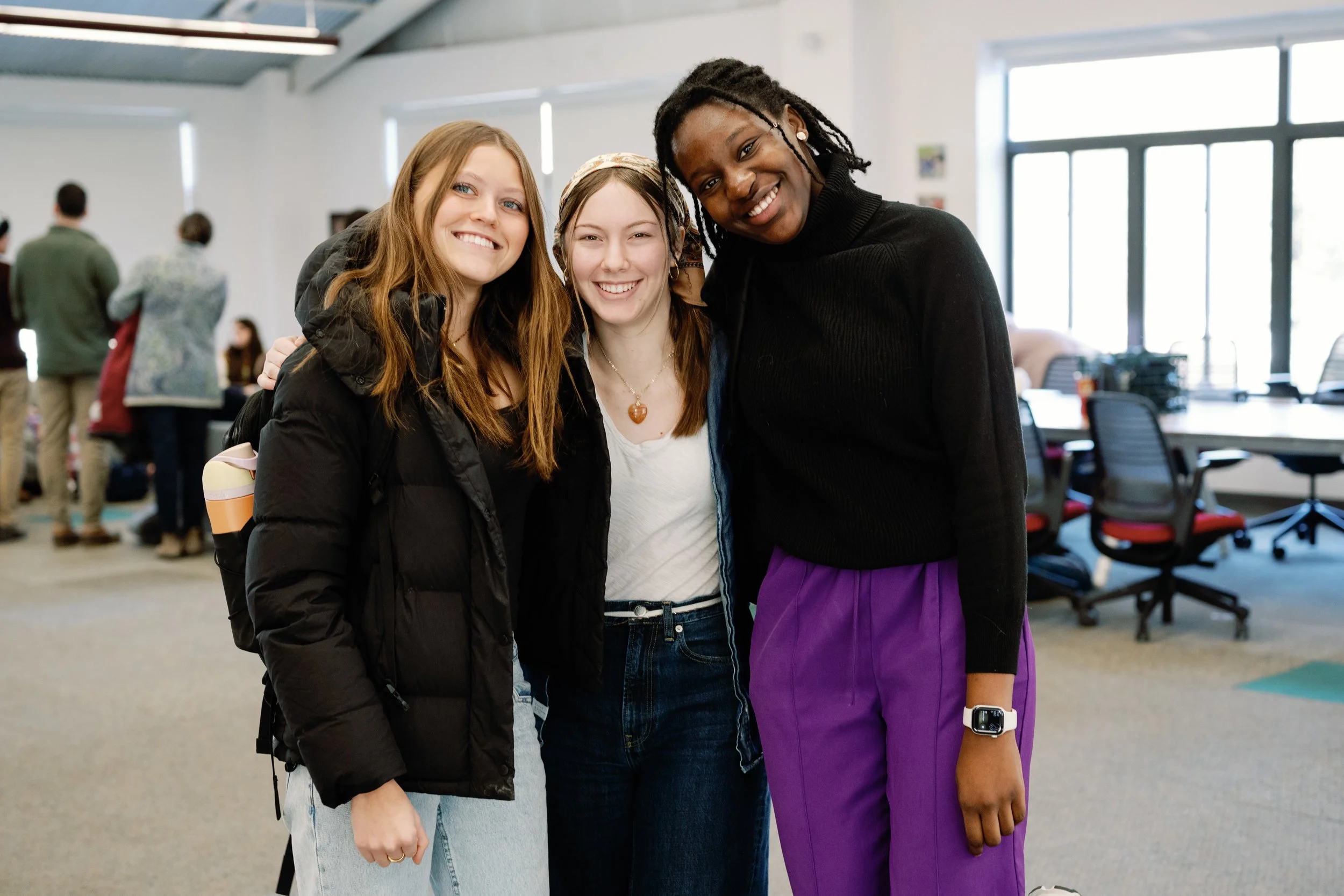 Three young women smiling and posing together in an indoor setting that resembles a classroom or meeting room with large windows and chairs in the background.