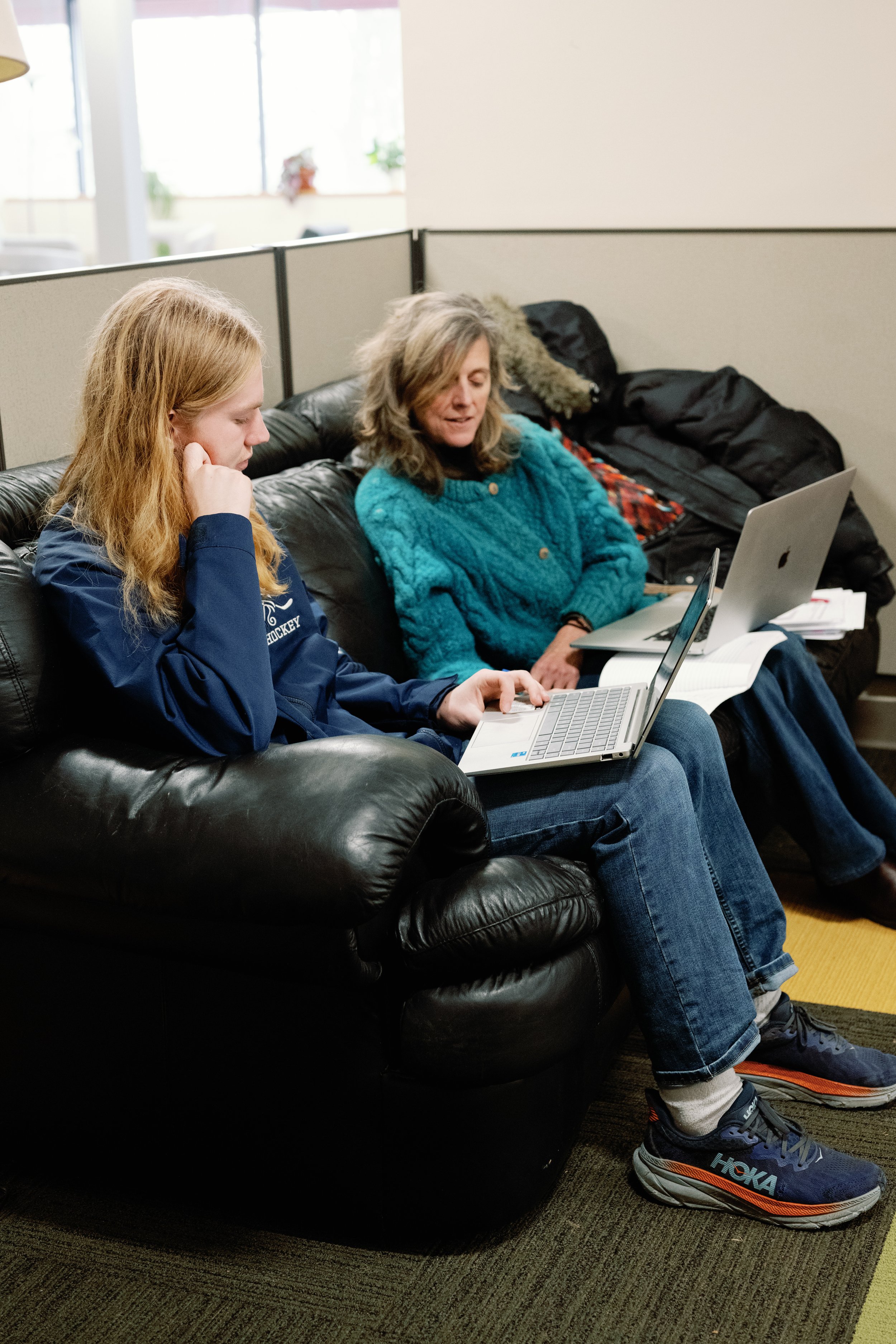 Two women seated on a black leather sofa using laptops, with papers and another laptop between them.