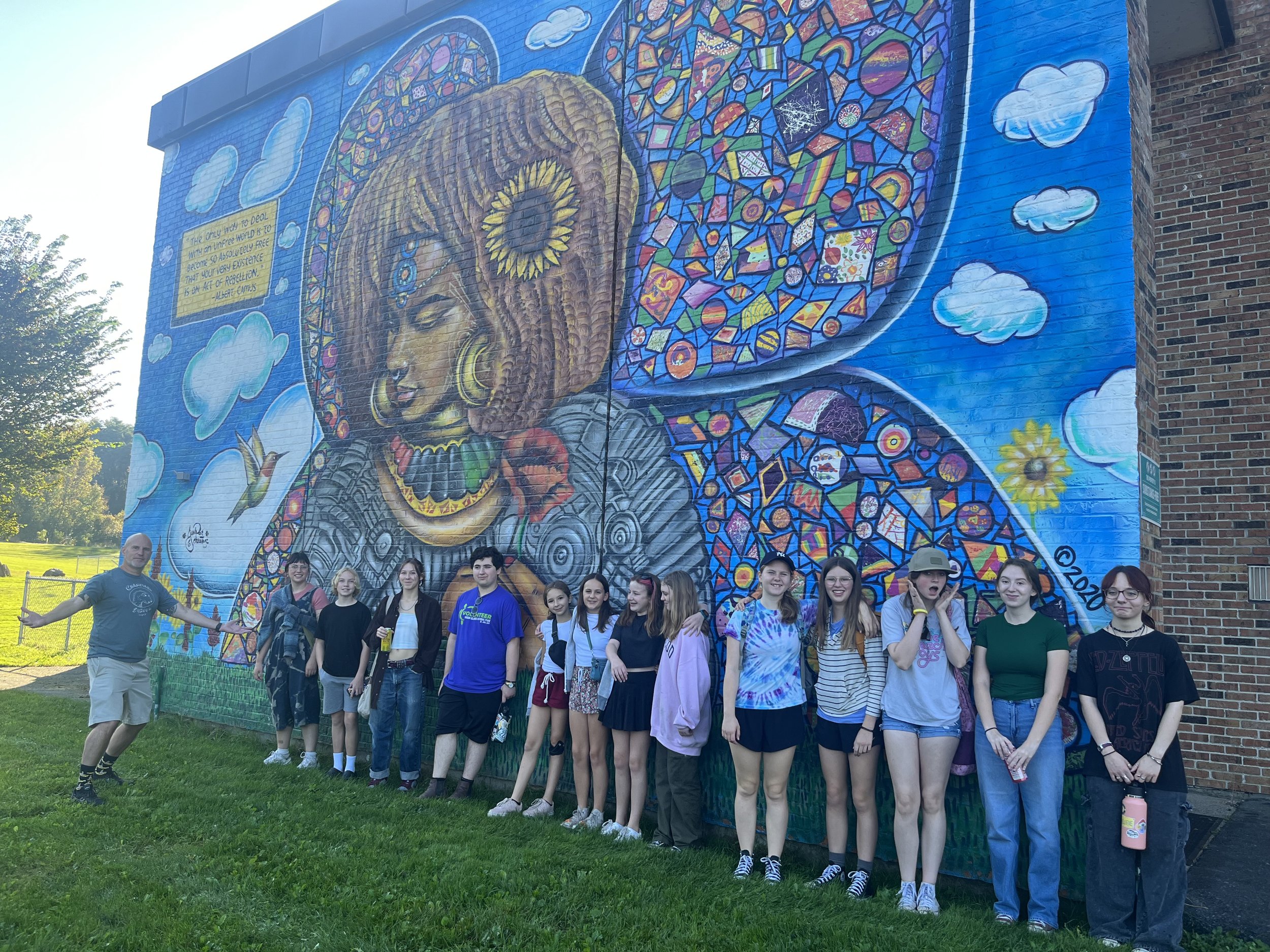 A group of people standing in front of a colorful mural on a brick building wall. The mural features a large woman's face with closed eyes, decorative hair, and vibrant patterns, clouds, a butterfly, and a sun. The group consists of children and adul