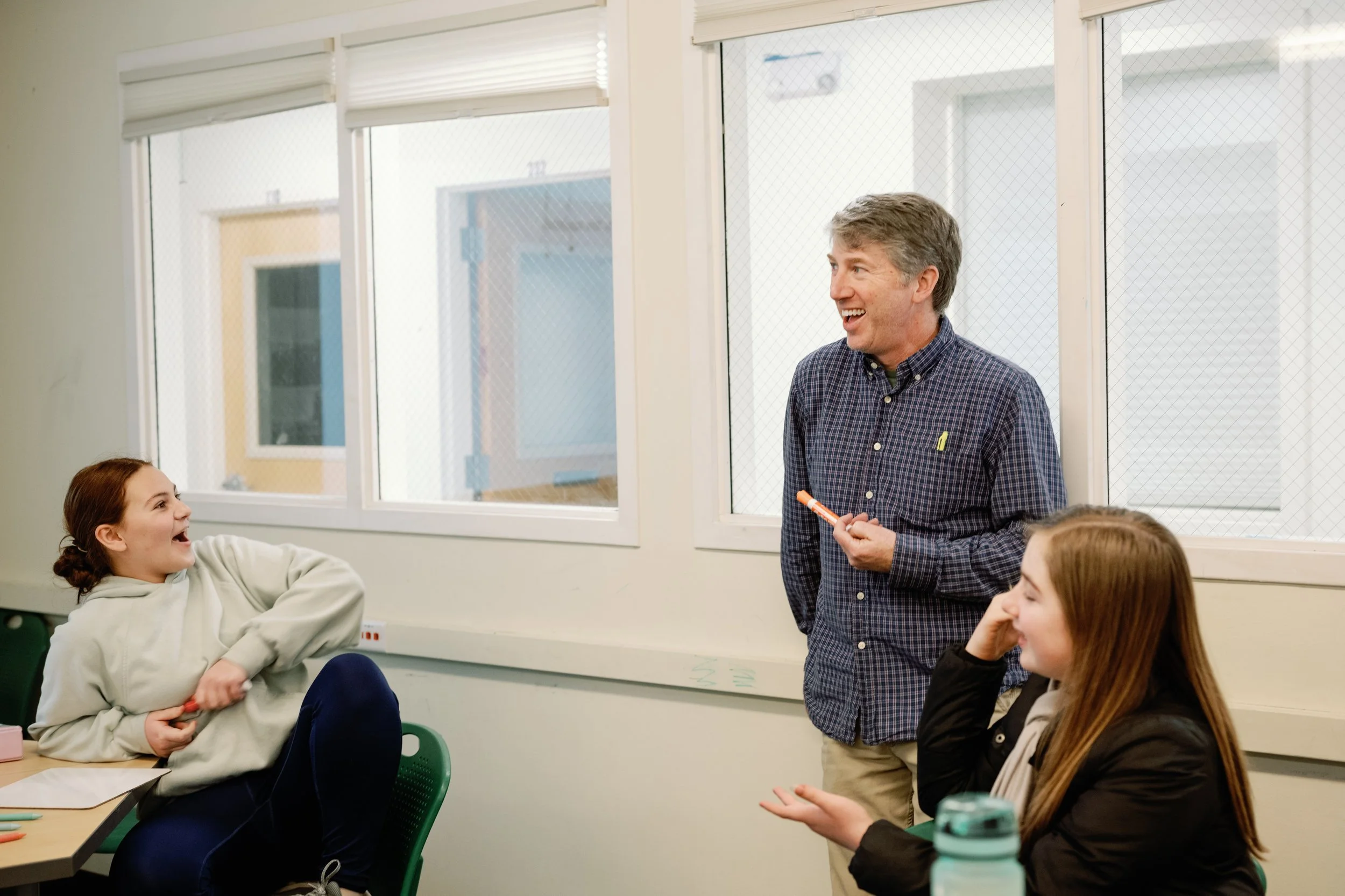 A classroom scene with two students and a teacher. The teacher is standing, talking, and holding a marker. Th  on the left, with red hair in a bun, is smiling and sitting at a desk. The girl on the right, with long brown hair, is listening and gestur