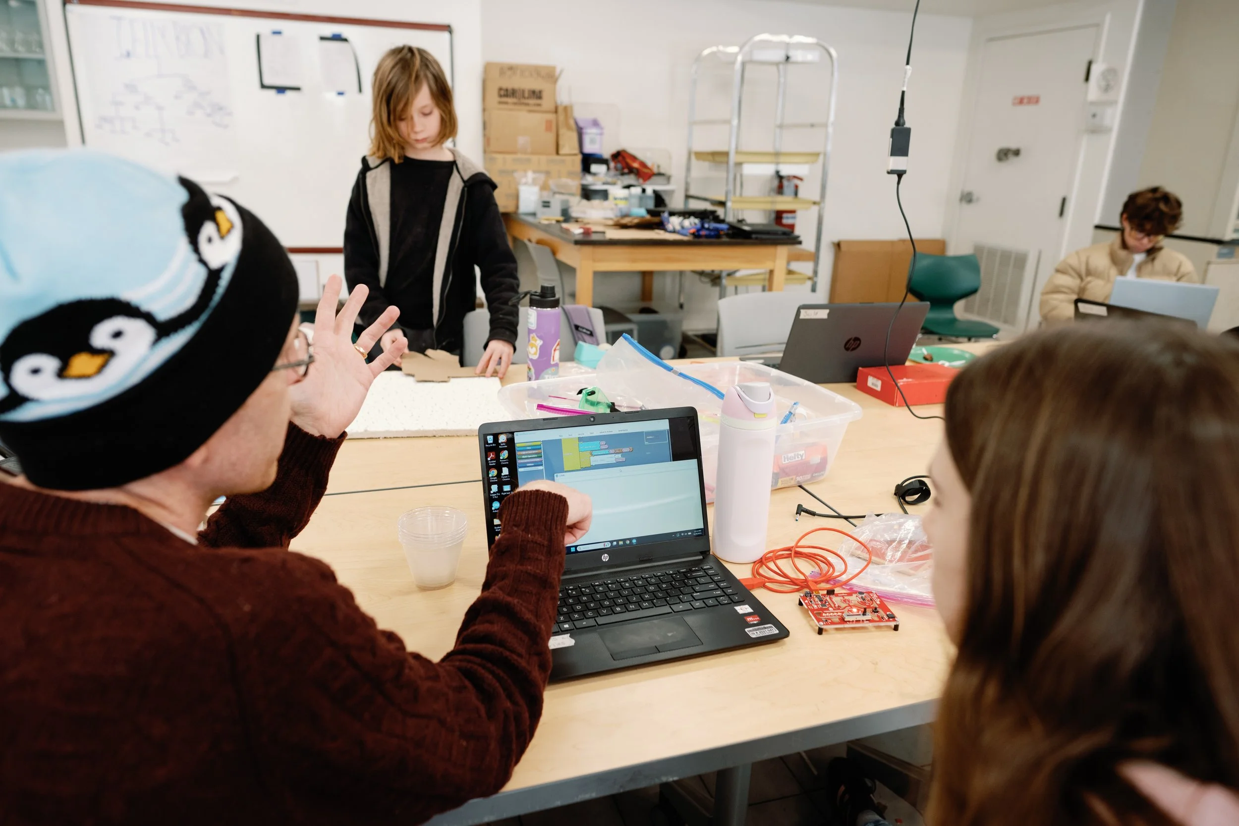 People gathered around a table working on a coding project with a laptop and electronic components in a classroom or workshop setting.