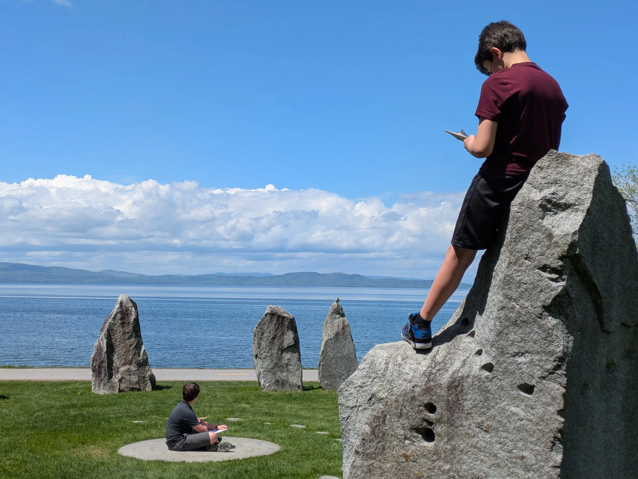 A boy in a maroon shirt and black shorts sitting on a concrete circle on grass, looking at a boy in a dark shirt and shorts standing on a large rock by a body of water with mountains and cloudy sky in the background.