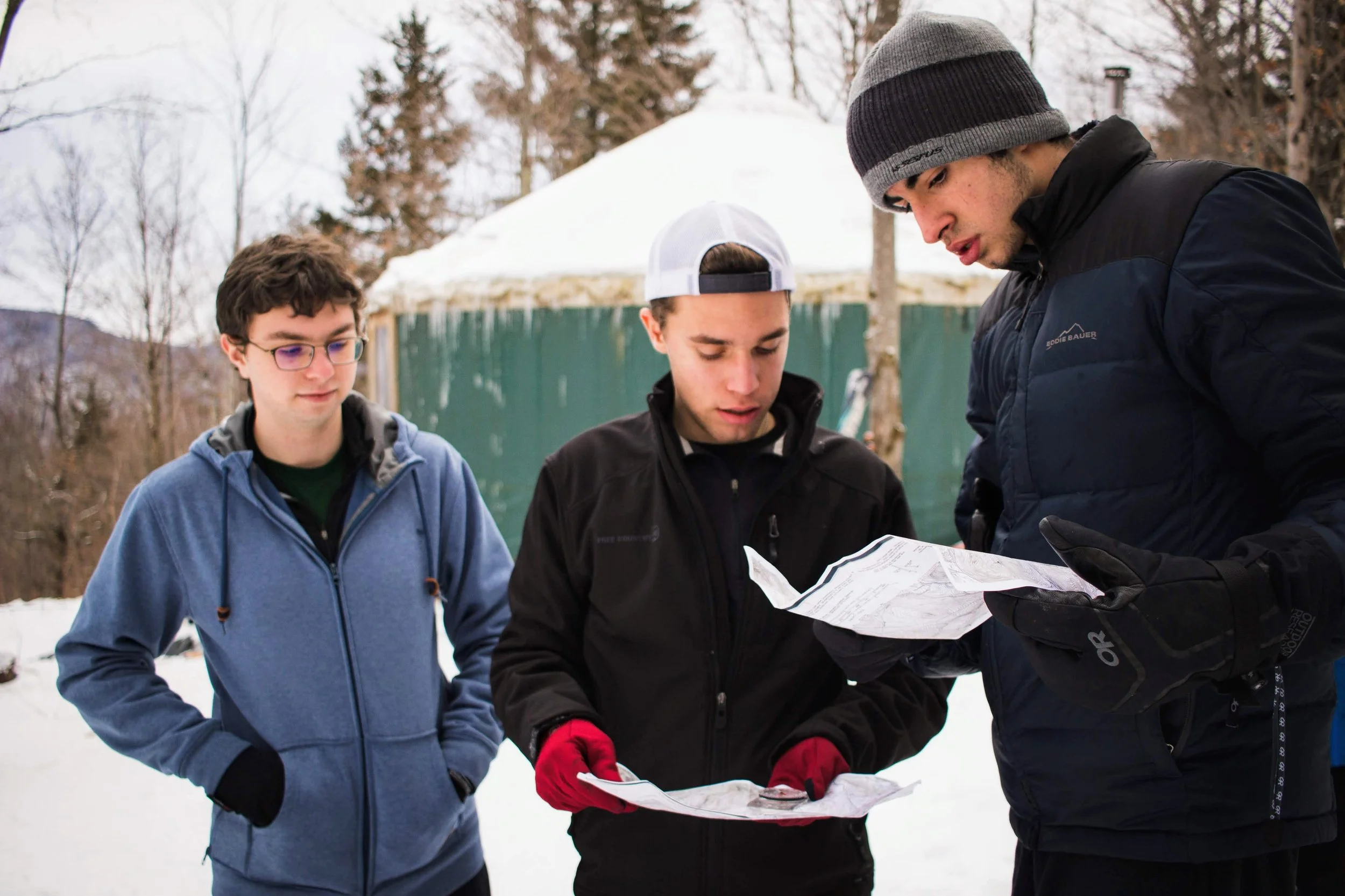 Three young men standing outdoors in a snowy area, examining a map together, with a snow-covered structure and trees in the background.