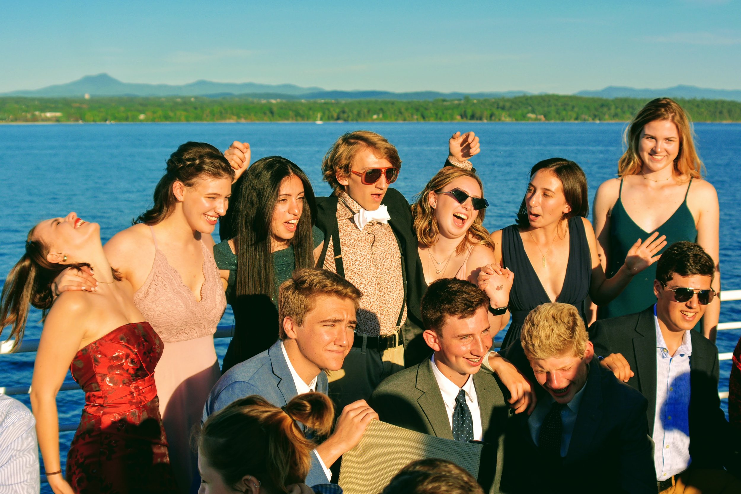 Group of young adults celebrating on a boat during daylight, with a water body and distant landscape in the background.