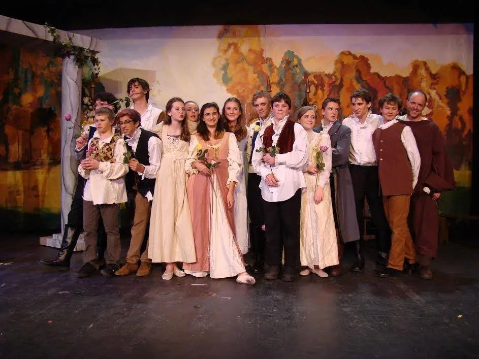 Group of young actors in period costumes on stage after a theatrical performance, holding roses, with a painted backdrop of colorful trees.