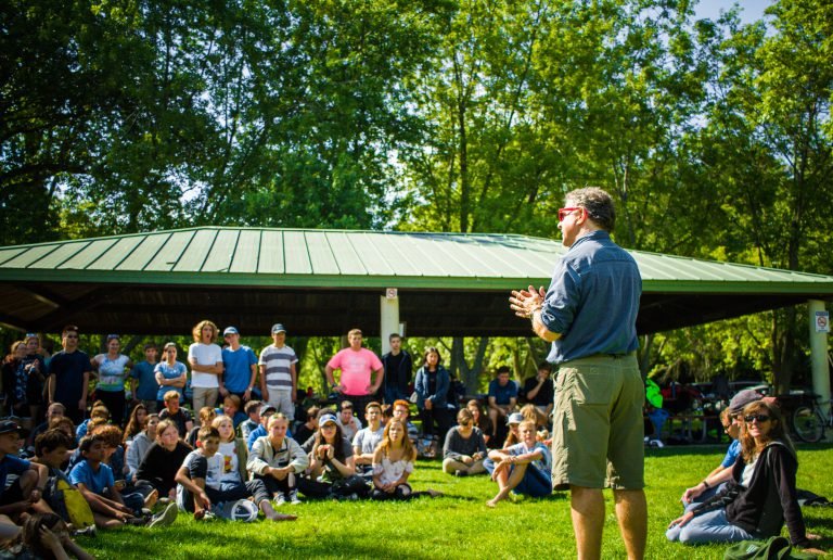 A man speaking to a large crowd of people gathered outdoors in a park, with a shelter and trees in the background.