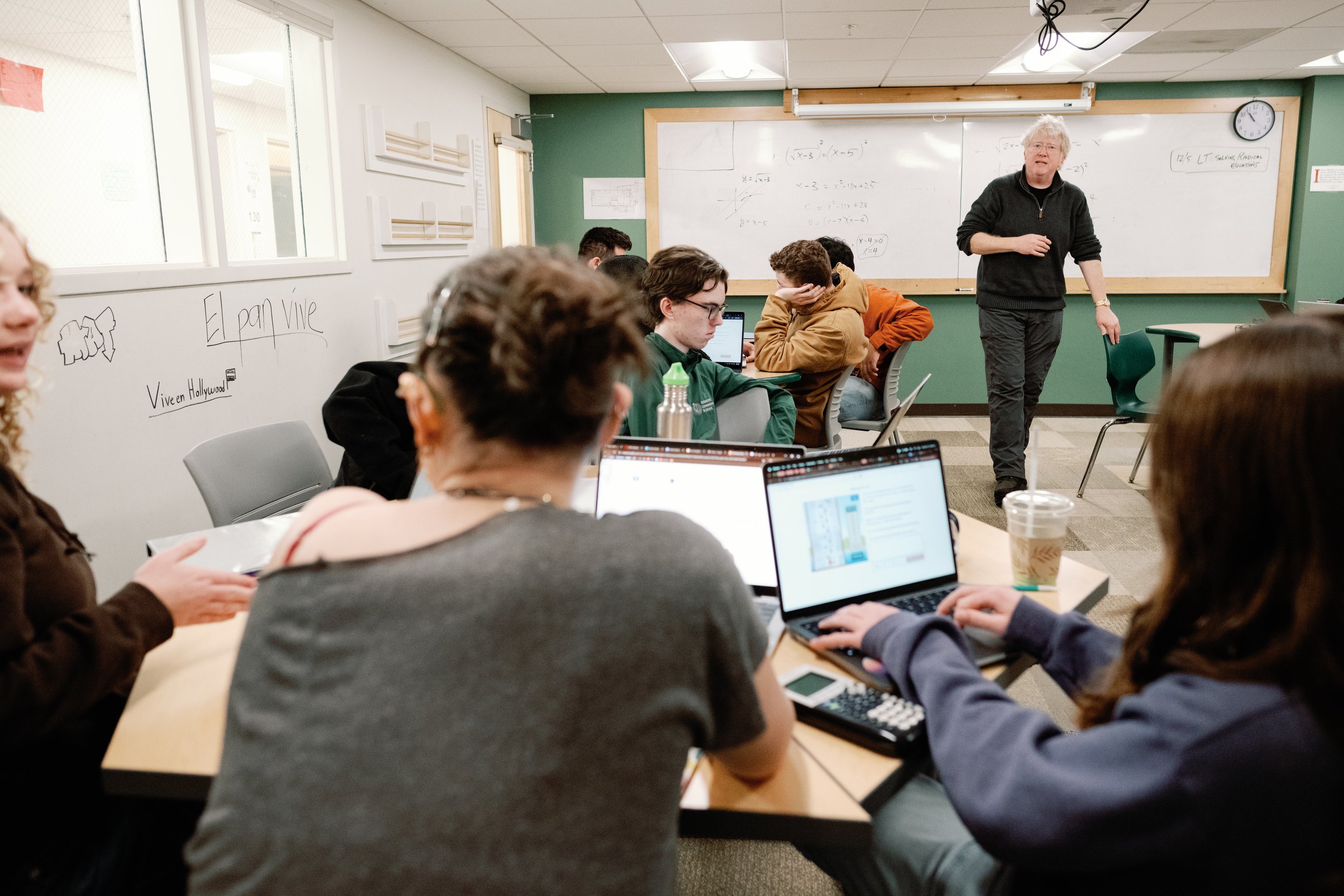 A classroom scene with students taking notes and using laptops while a teacher explains algebra problems on the whiteboard.