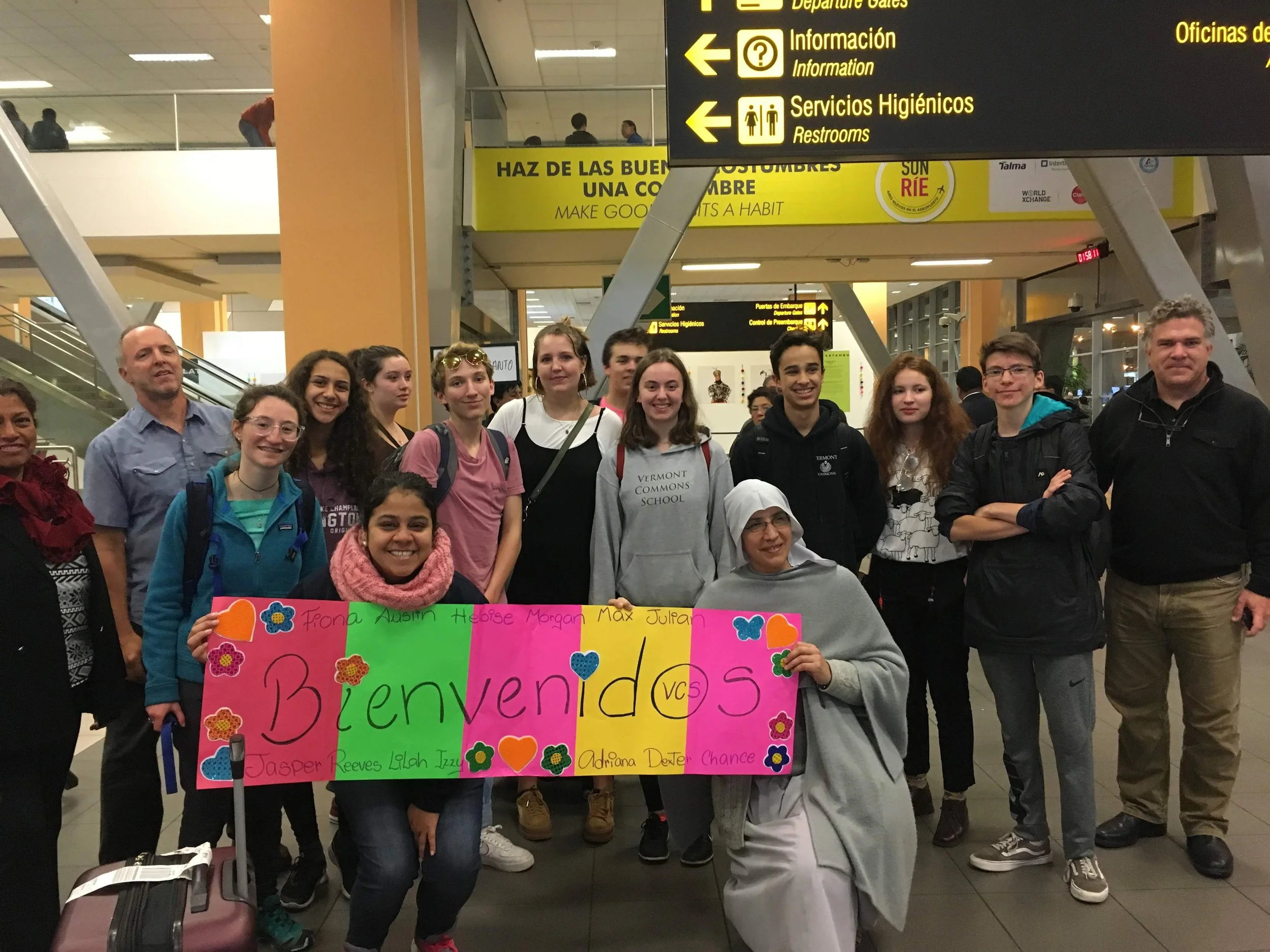 Group of young people and adults holding a colorful welcome sign in an airport, with signs indicating gates, information, restrooms, and services overhead.