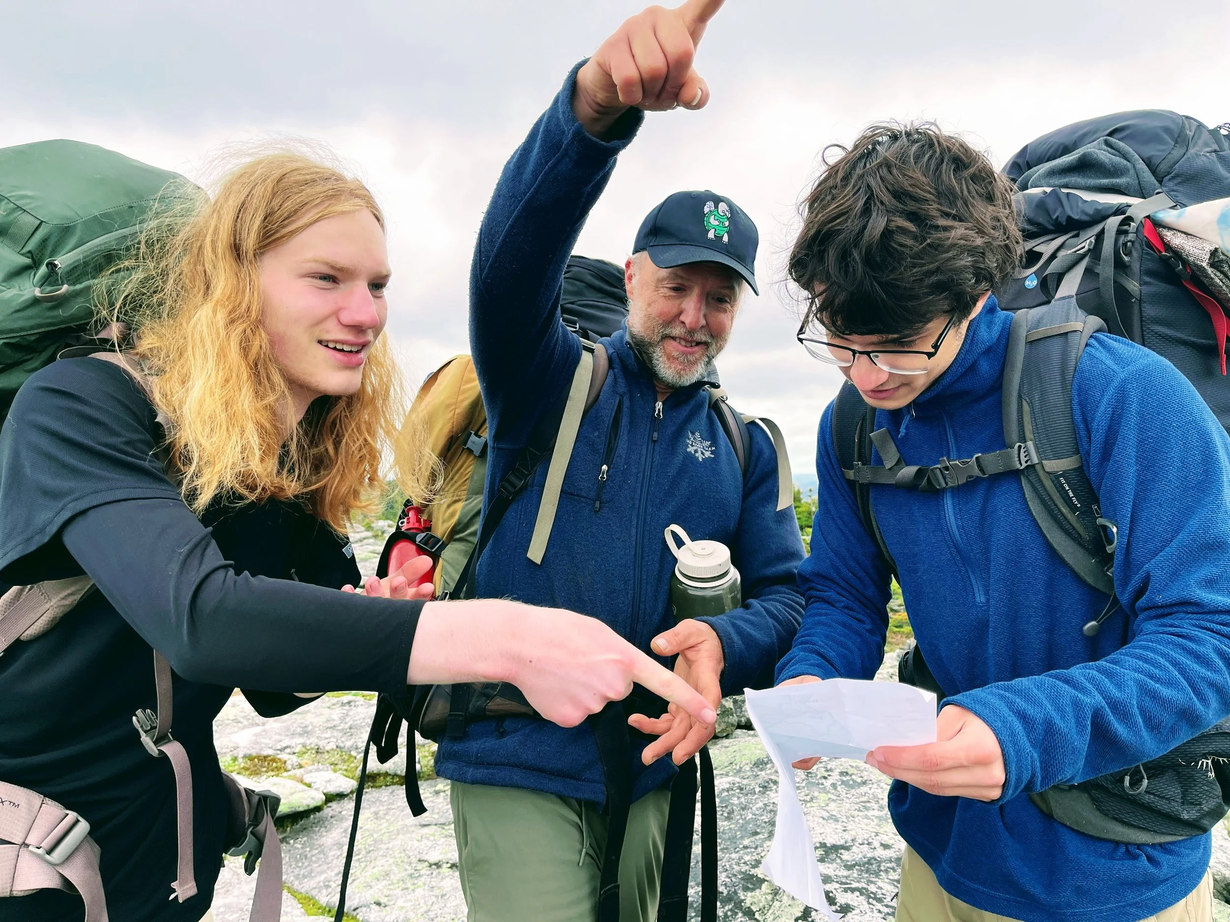 Three hikers with backpacks examining a map outdoors on a cloudy day.