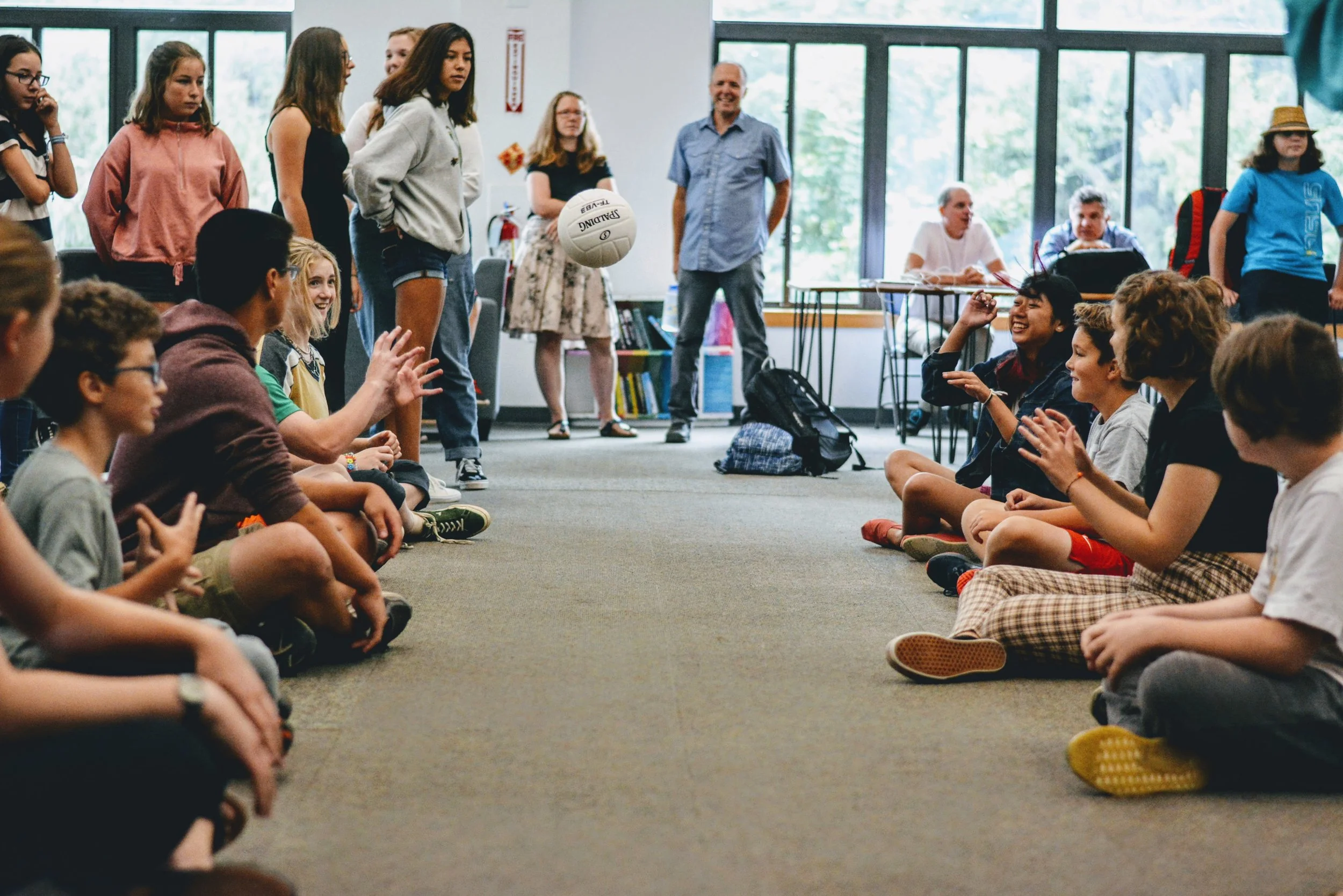 Group of children and adults in a room, with some kids sitting on the floor facing each other, and a girl in the middle holding a volleyball. The room has large windows, and some adults are standing or sitting at tables in the background.