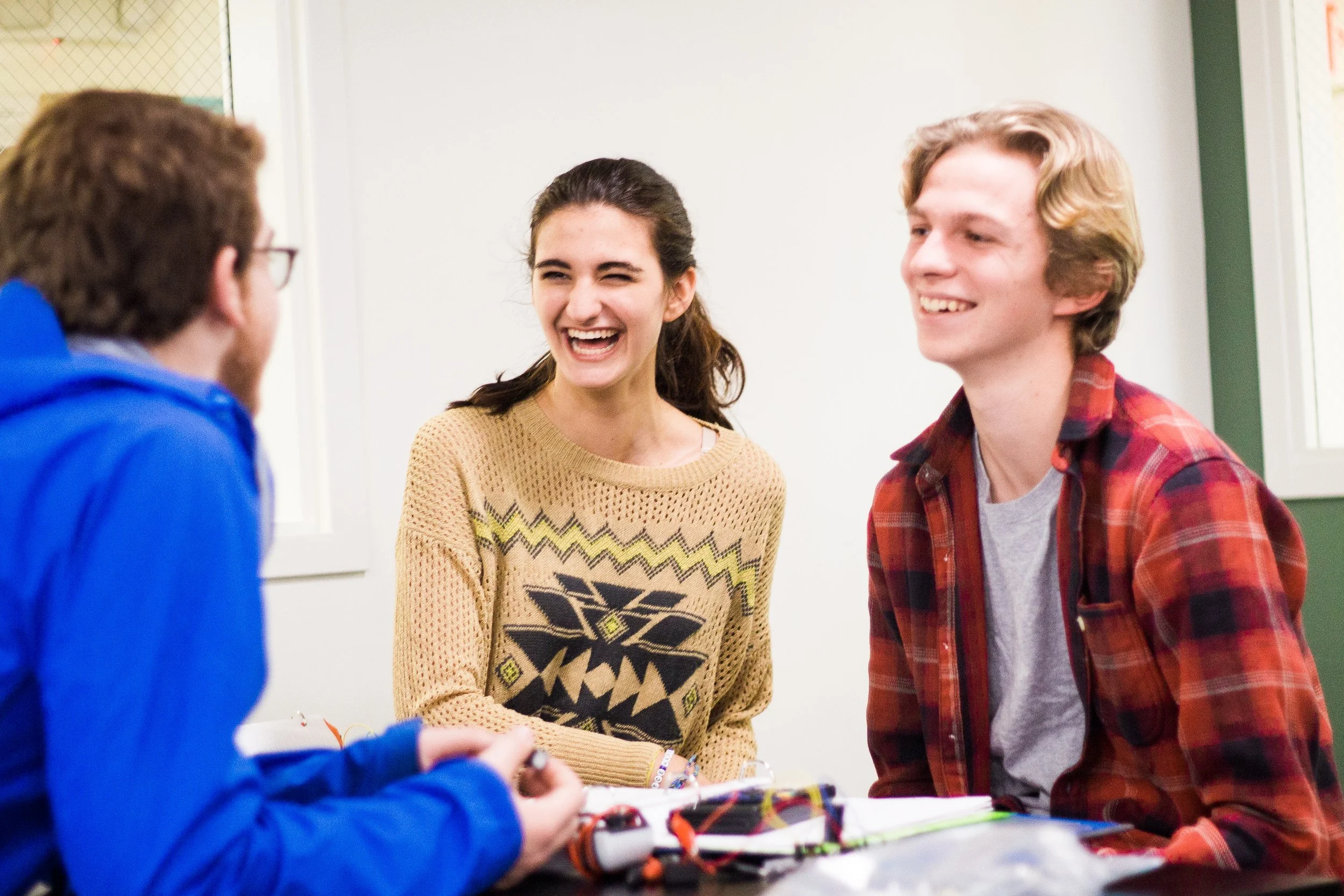 Three young people laughing and talking at a table with electronic components and wires.