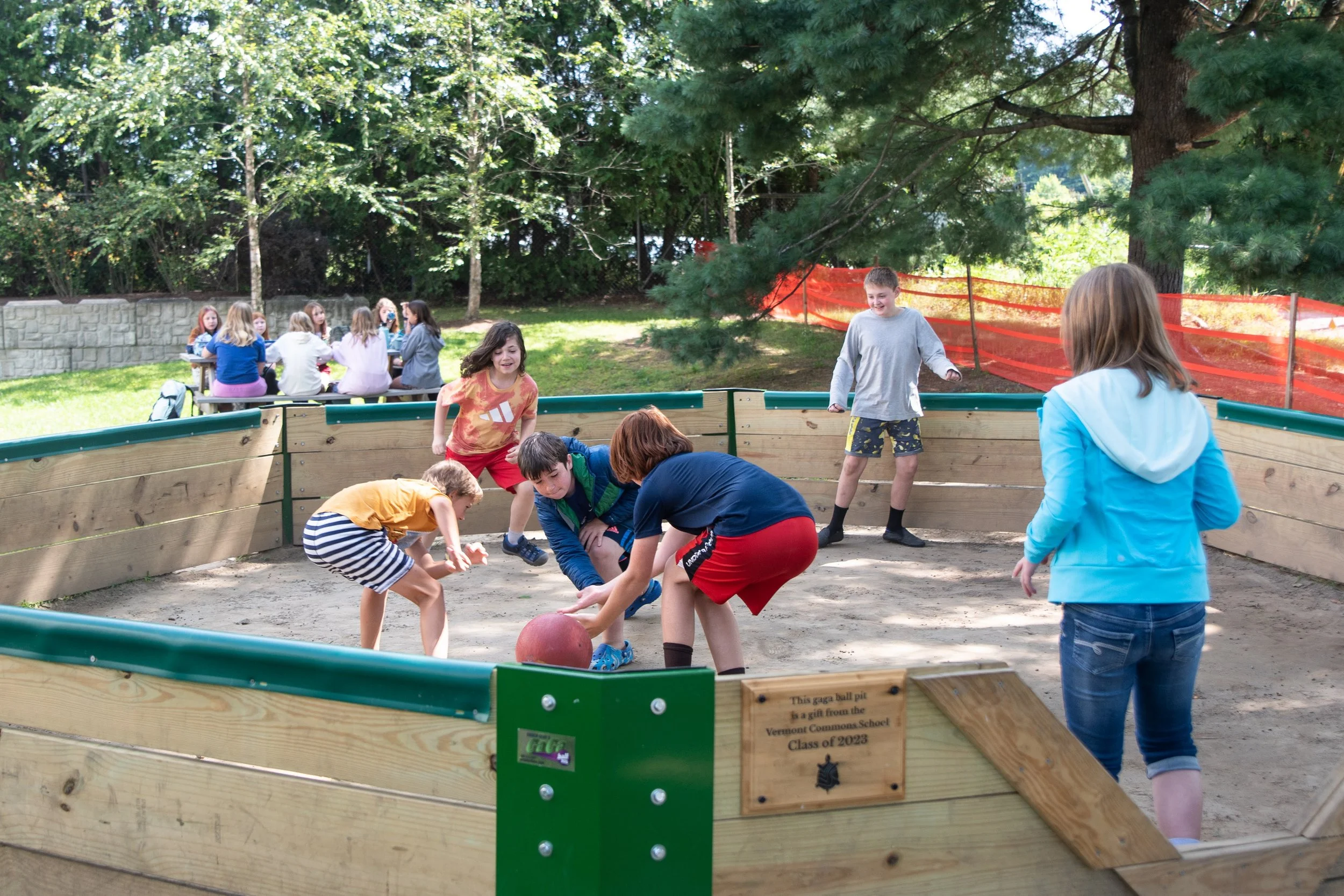 Children playing a game of gaga ball in a wooden, outdoor pit surrounded by trees and other kids sitting at a table in the background.