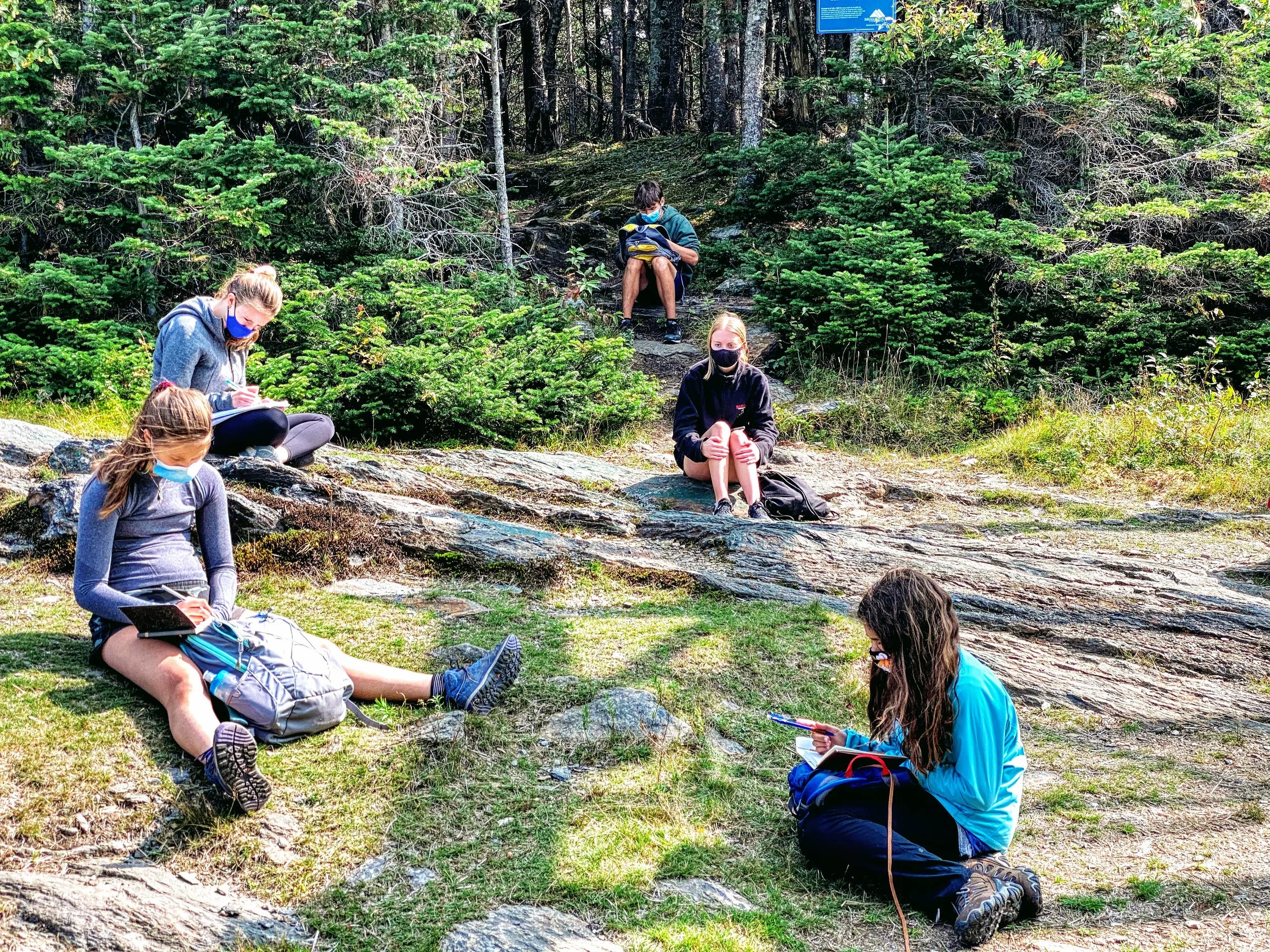 Six young people sitting outdoors in a forested area, some taking notes or reading, all wearing masks and casual clothing, and surrounded by green trees and rocks.