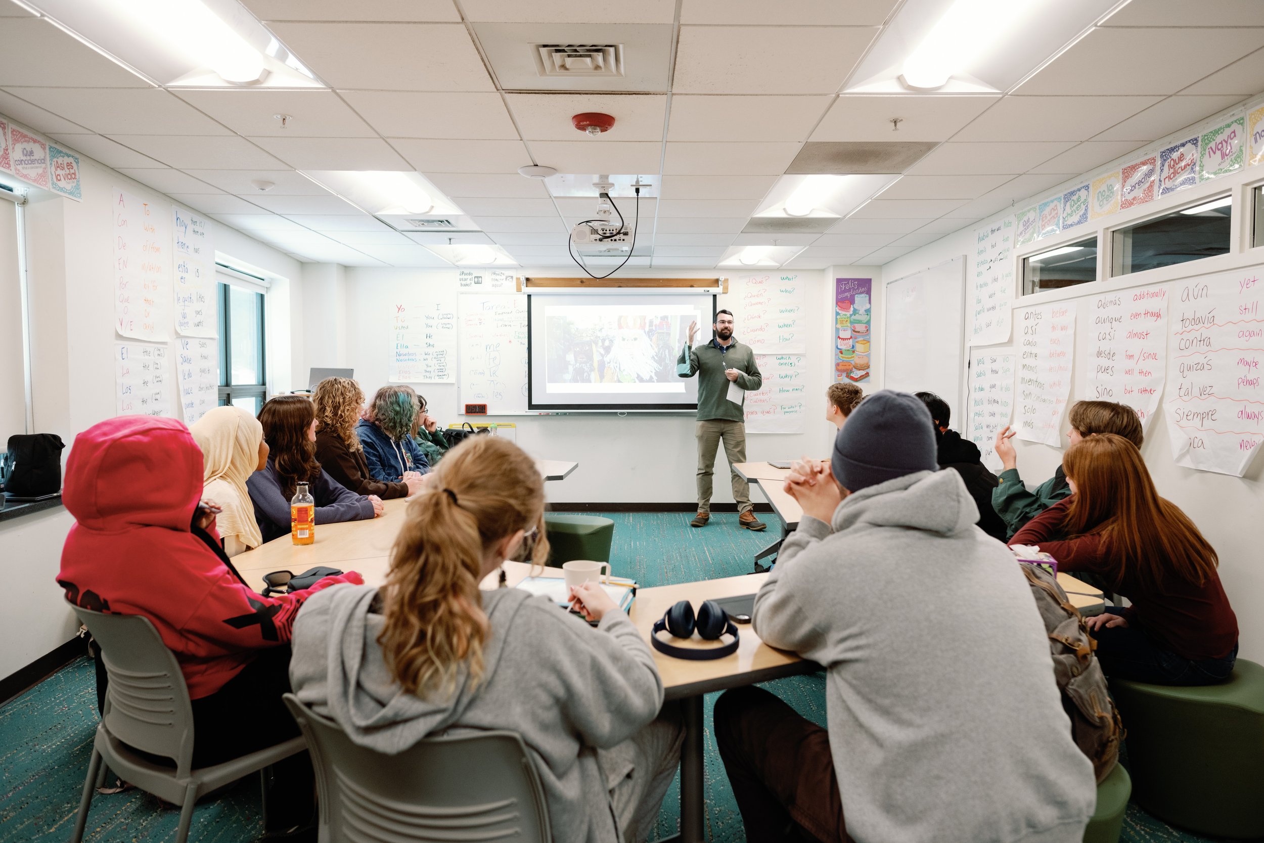 A classroom with students sitting around a U-shaped table, listening to a male instructor at the front. The instructor is gesturing towards a projection screen with trees. The classroom walls are decorated with posters and large sheets of paper with writings.