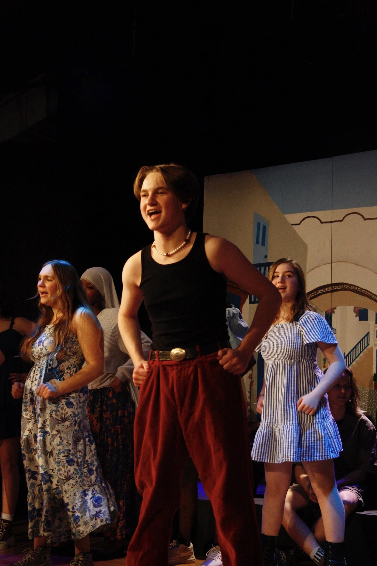 Young women performing on stage, dressed in casual attire, with a painted background of a town scene.