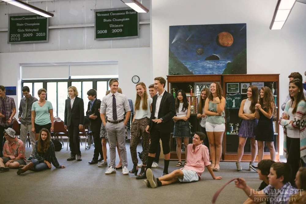Group of students gathered inside a school gymnasium, some standing and some sitting on the floor, engaging in a casual activity or presentation. School banners hang from the ceiling, and a large space-themed painting is visible on the wall.