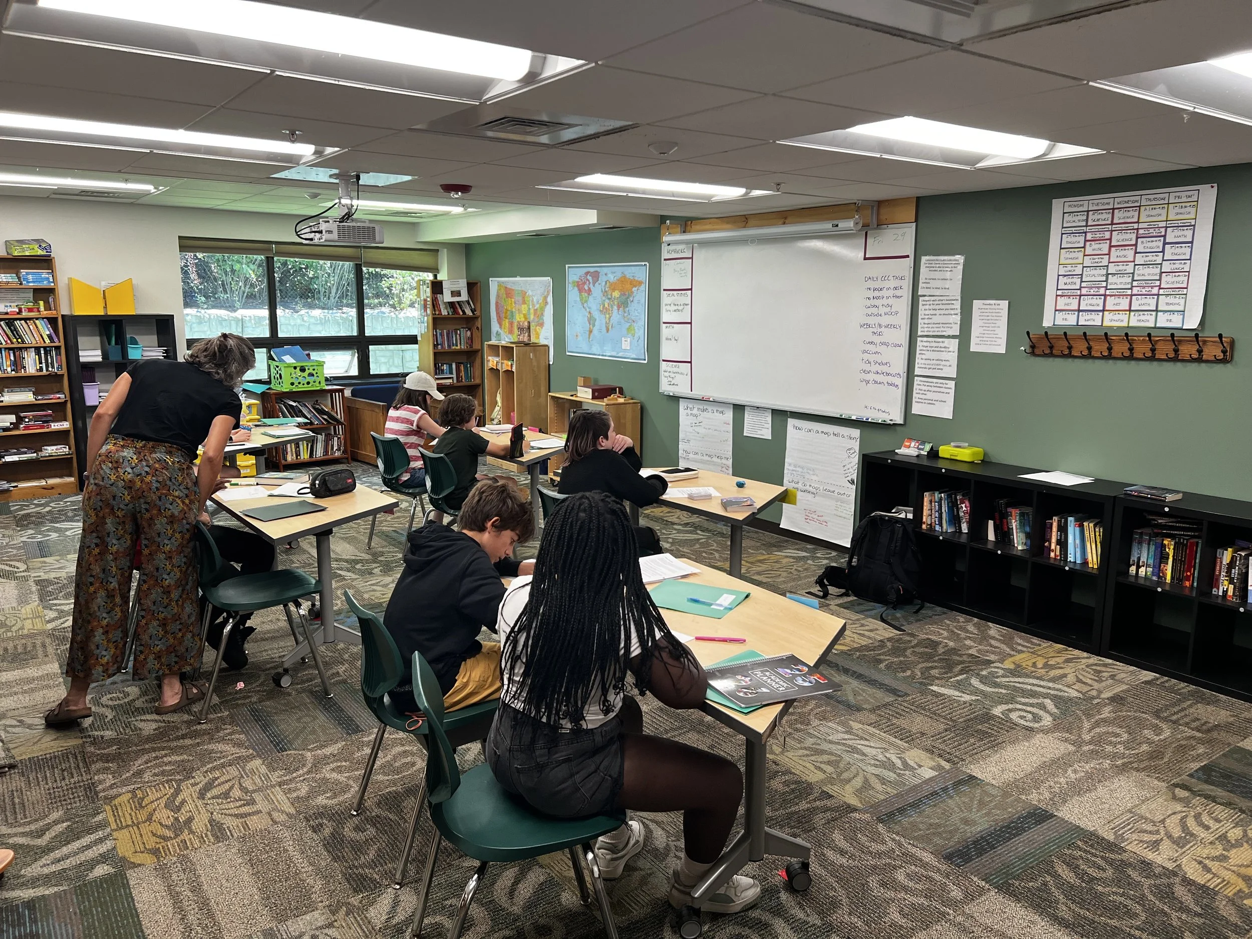 A classroom with students seated at desks, a teacher standing, and educational materials on the walls and shelves.