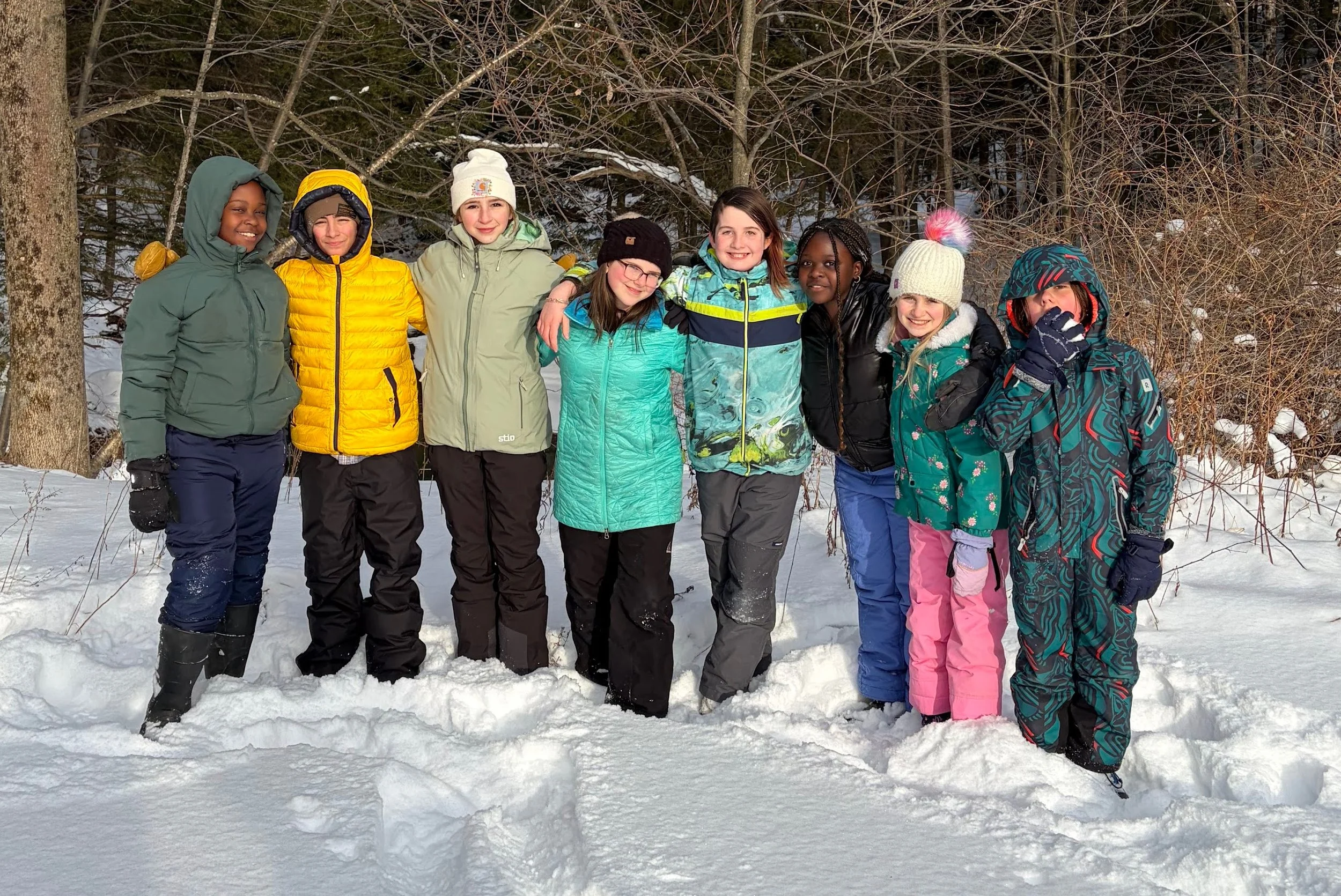 A group of eight children dressed in winter clothing standing in the snow outdoors, smiling and posing for the photo with trees in the background.
