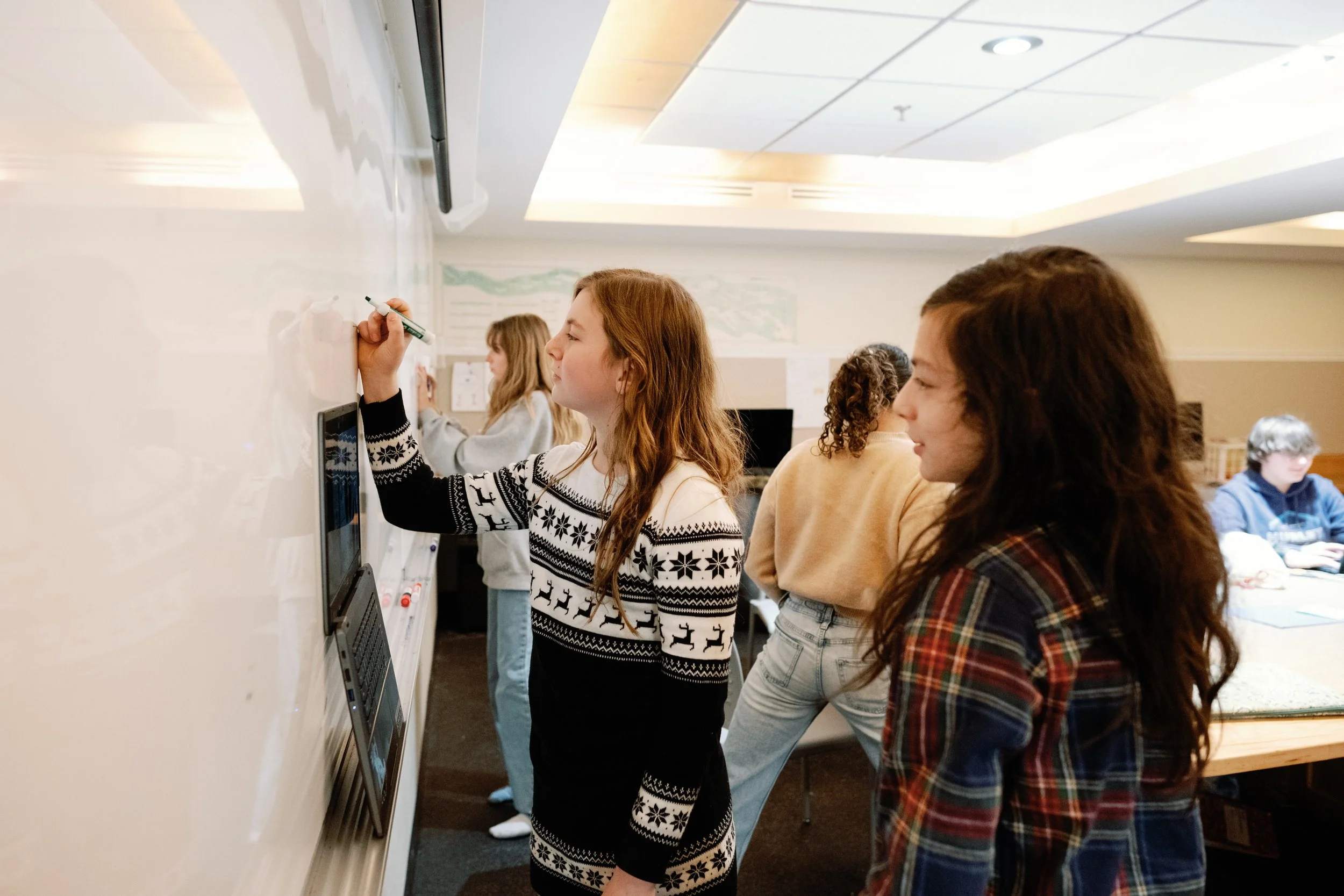Girls in a classroom writing on a whiteboard, with others seated at tables in the background.
