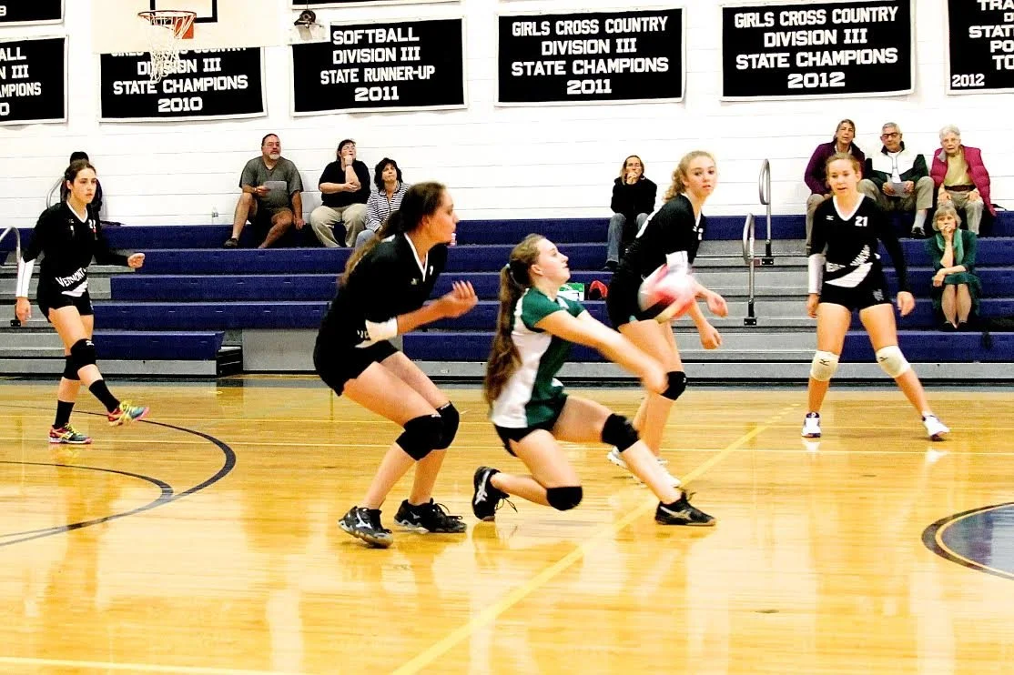 Girls playing volleyball on an indoor court with spectators in the background and championship banners hanging on the wall.