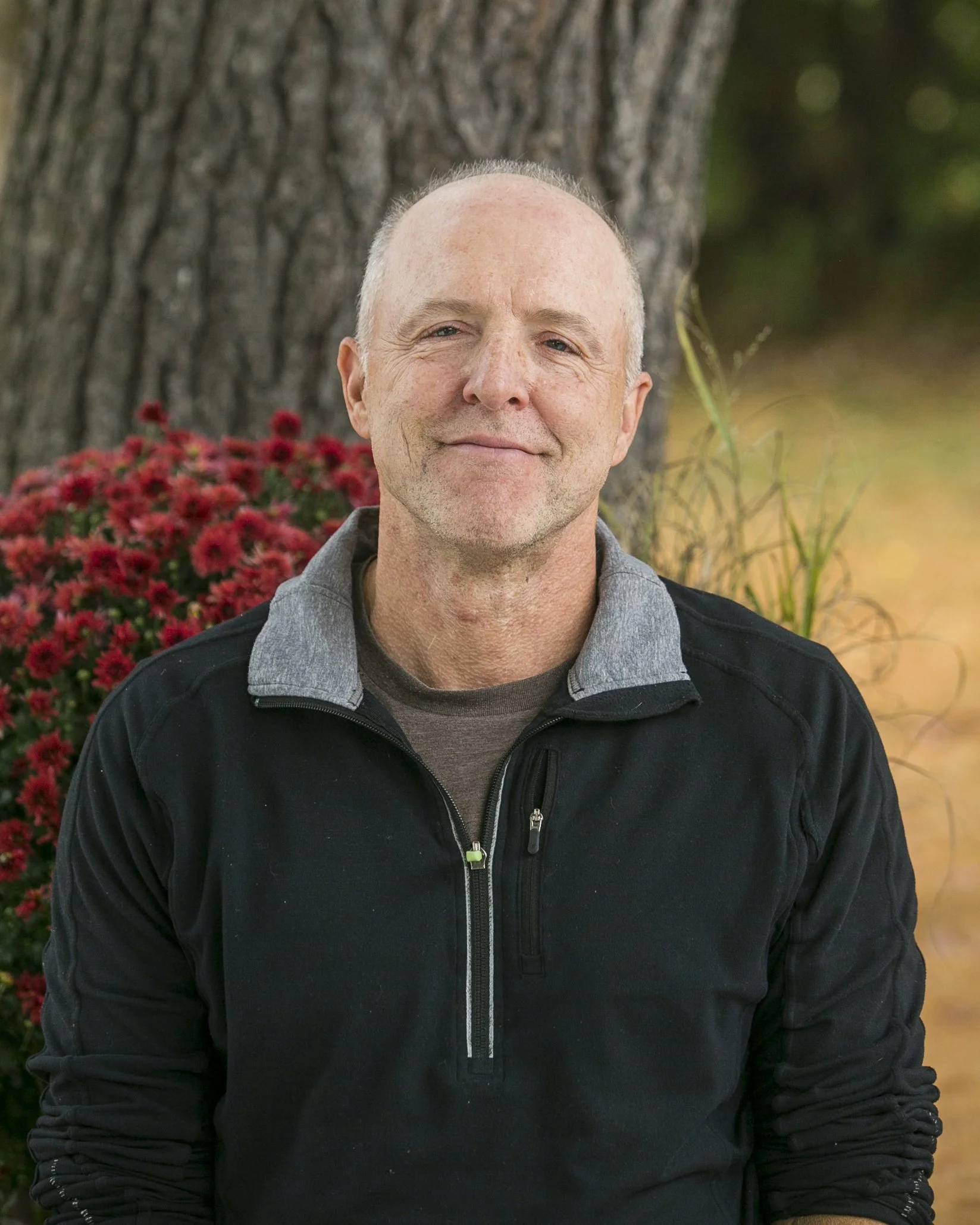 A man with a slight smile, no shirt, seated outdoors with a tree and red flowers behind him.