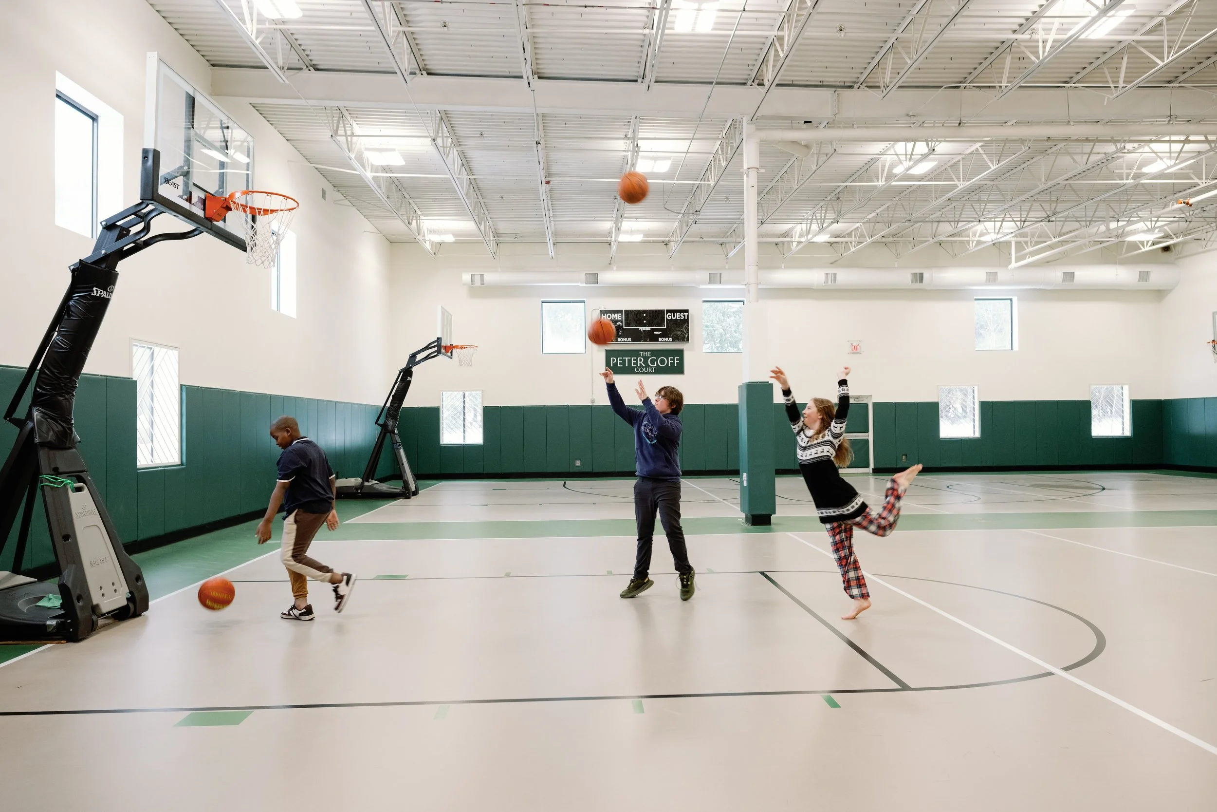 Children playing basketball indoors at a gymnasium, with two kids shooting hoops and a third kid playing on the floor.