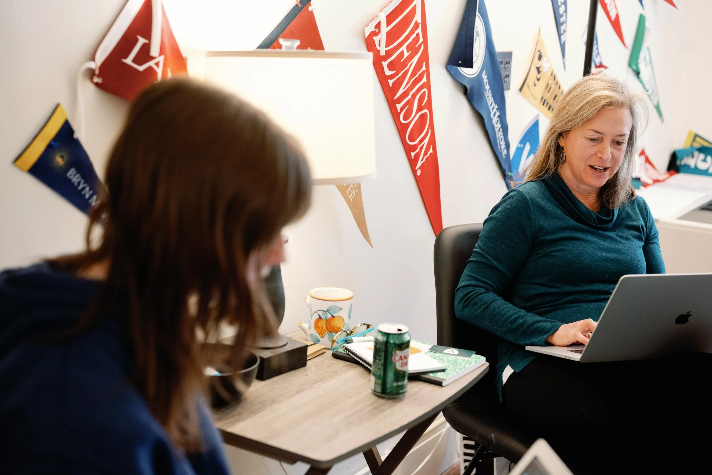 Two women sitting at a table in an office decorated with colorful college pennants on the wall. One woman is looking at a laptop, the other is facing away from the camera.
