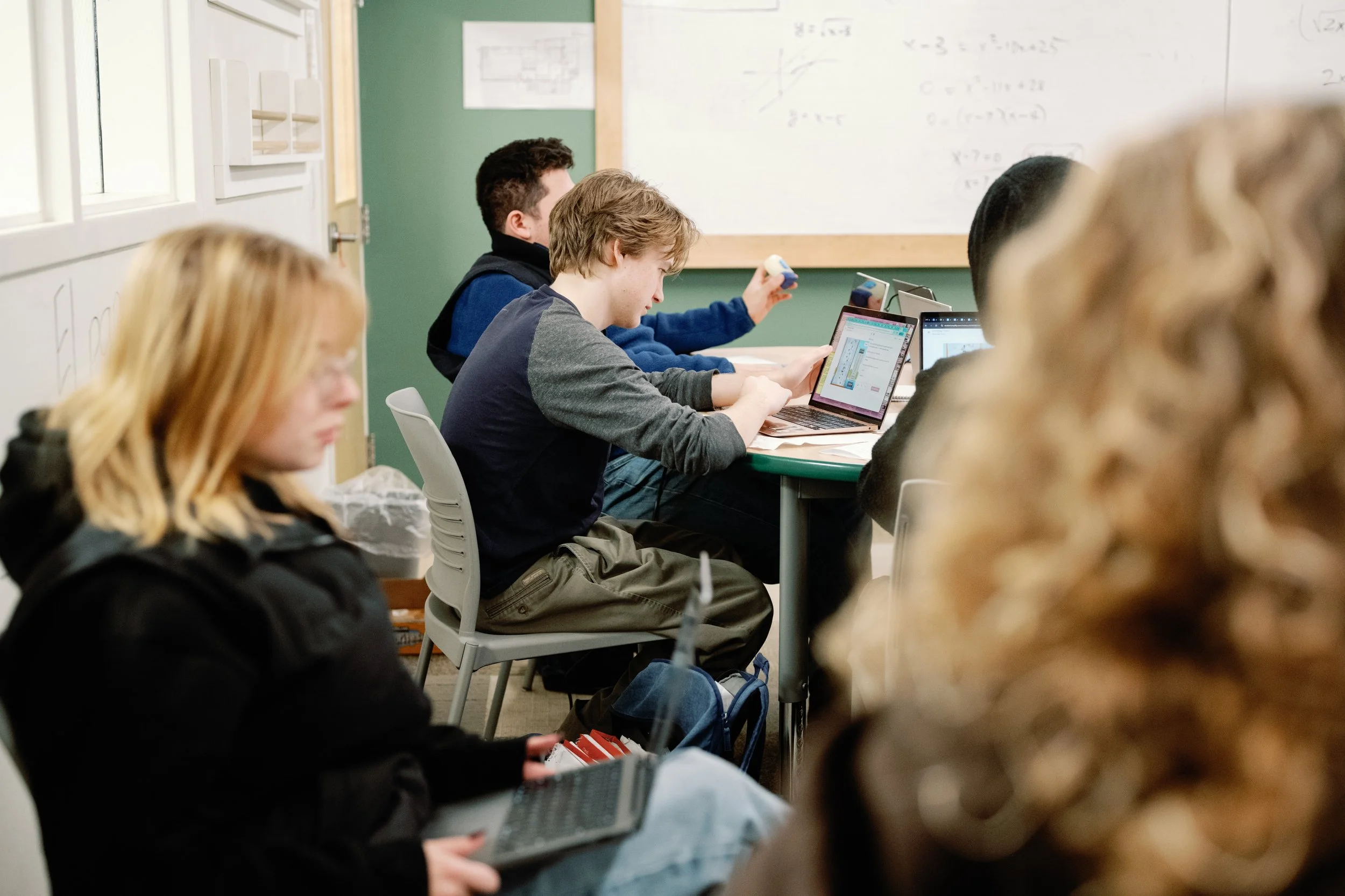 A classroom with students using laptops and tablets, with a whiteboard in the background containing mathematical equations.