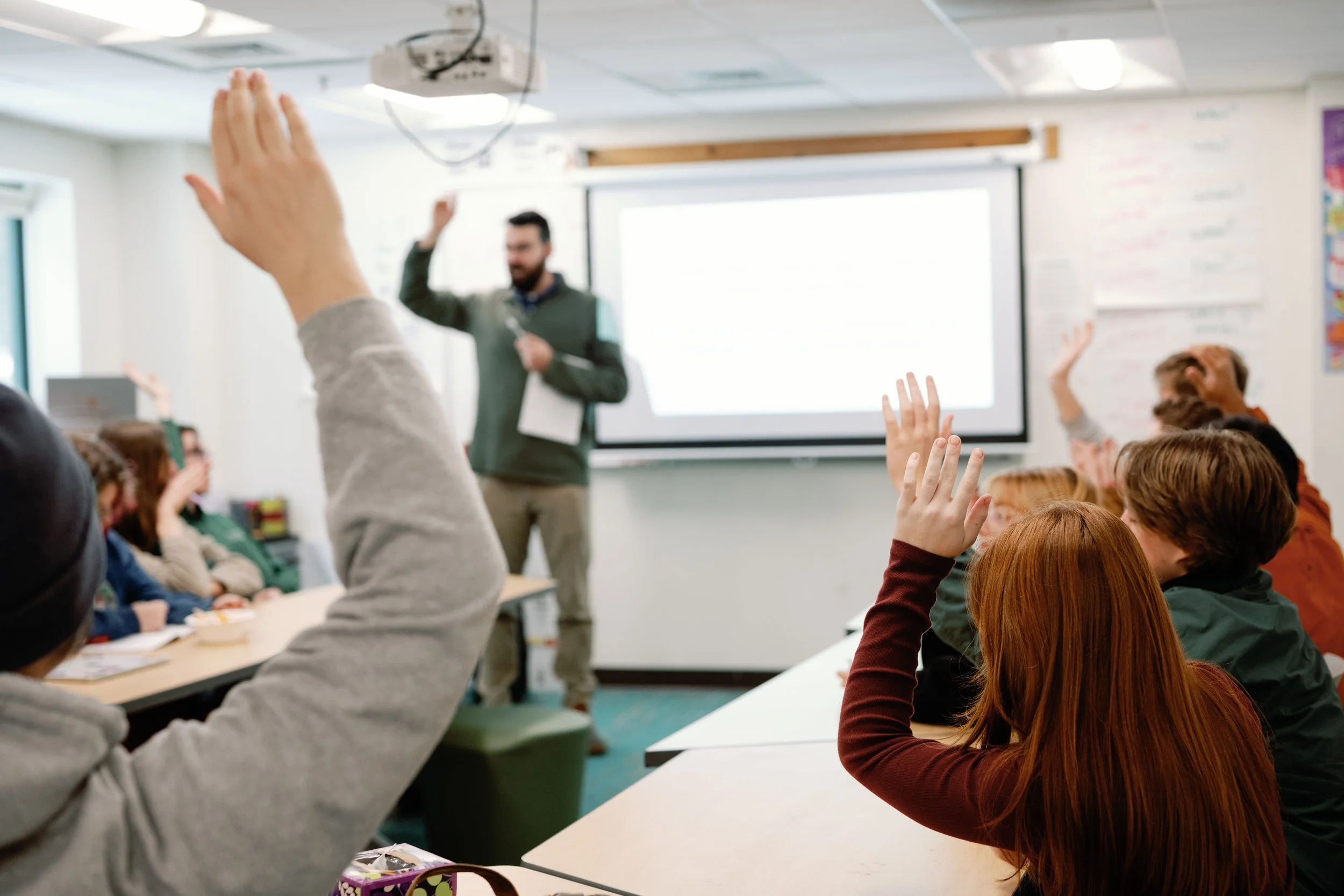 Classroom with students raising hands and a teacher at the front near a projection screen