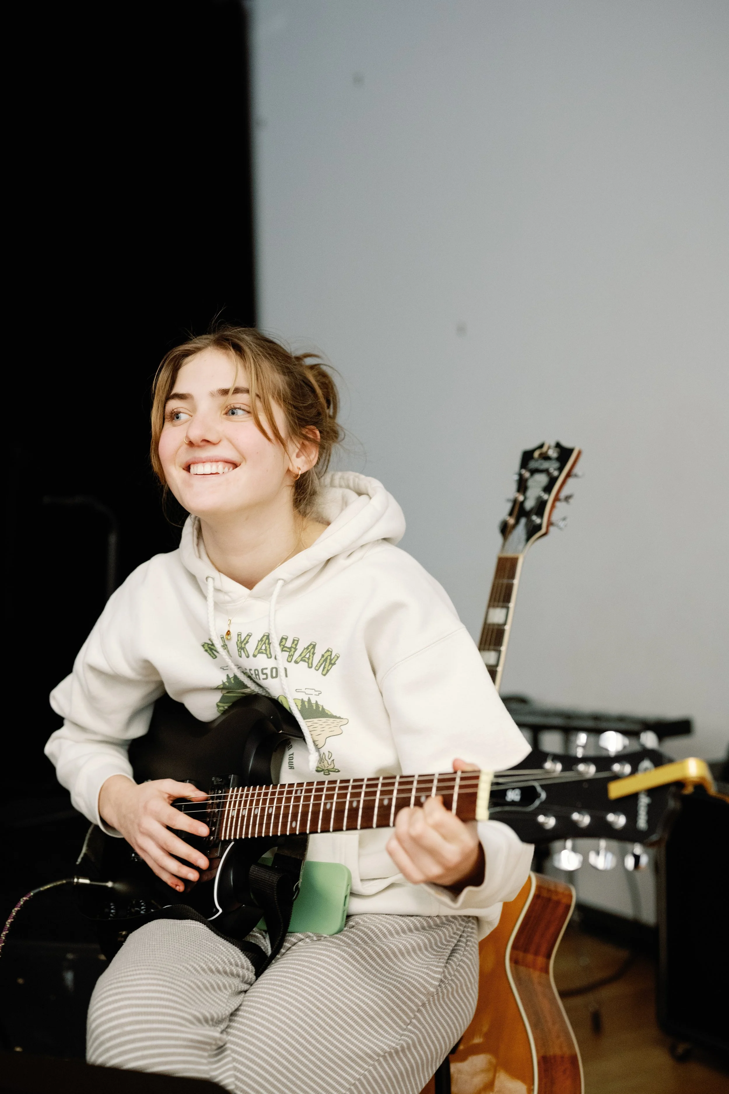 A young woman with light brown hair smiling while playing an electric guitar in a music rehearsal space.