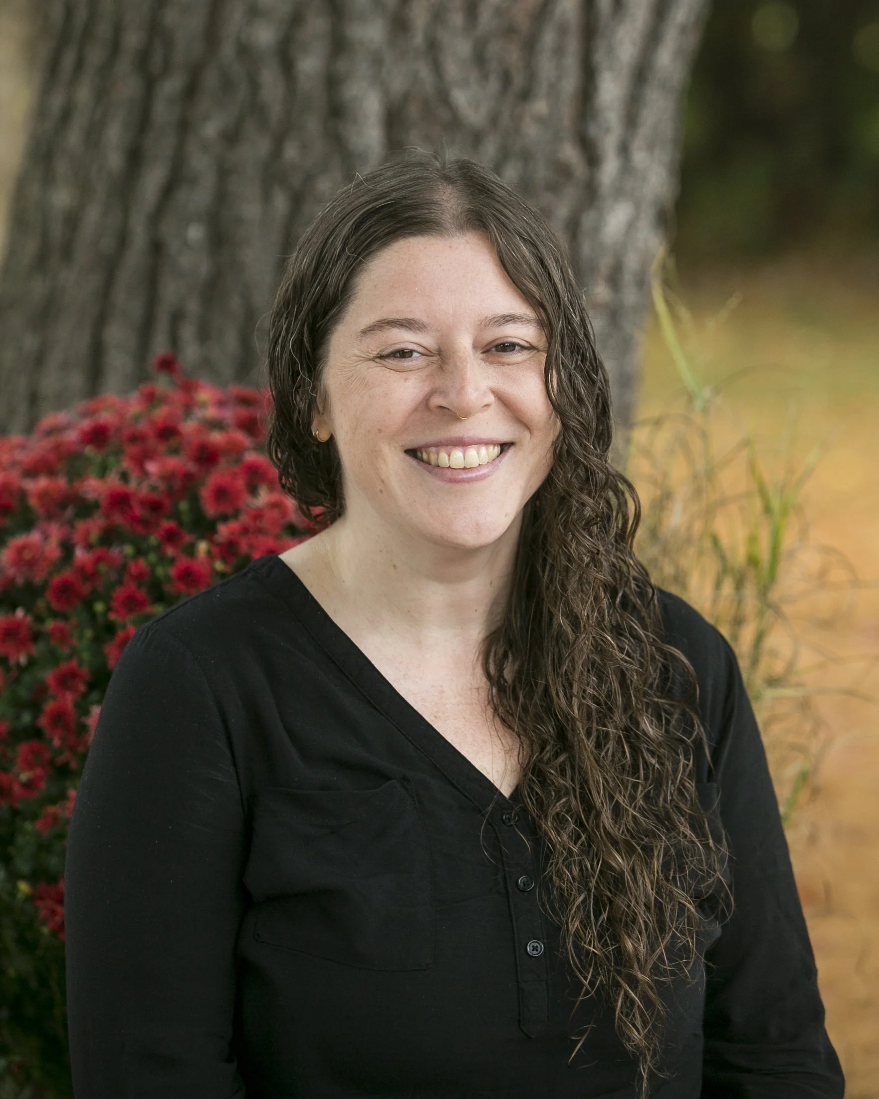 A woman with long, curly brown hair, wearing a black top, smiling, standing outdoors against a background of a tree trunk and red flowers.
