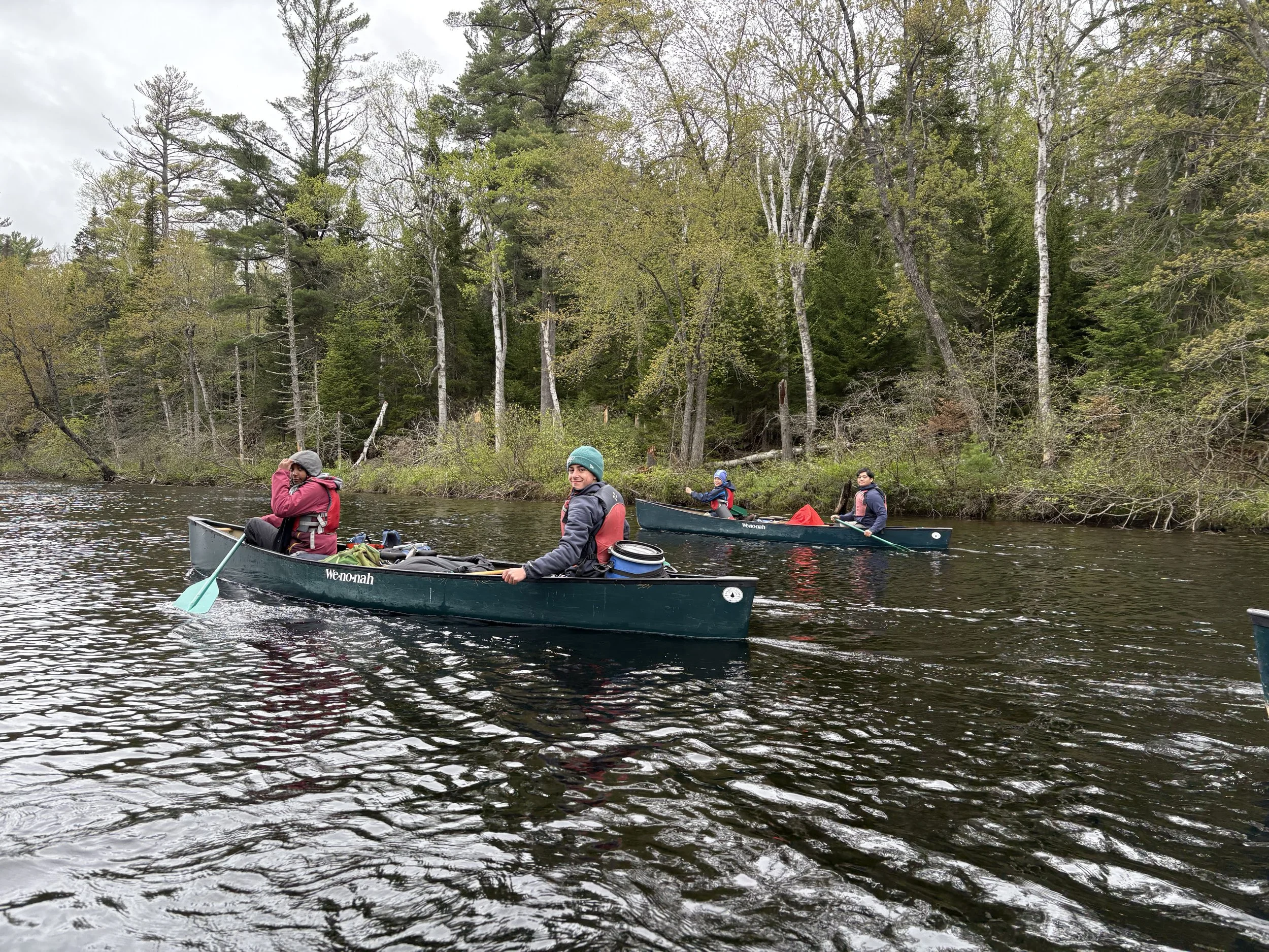 Four people canoeing on a river with a forested background, two in the foreground and two in the background, all wearing life jackets and jackets for weather.
