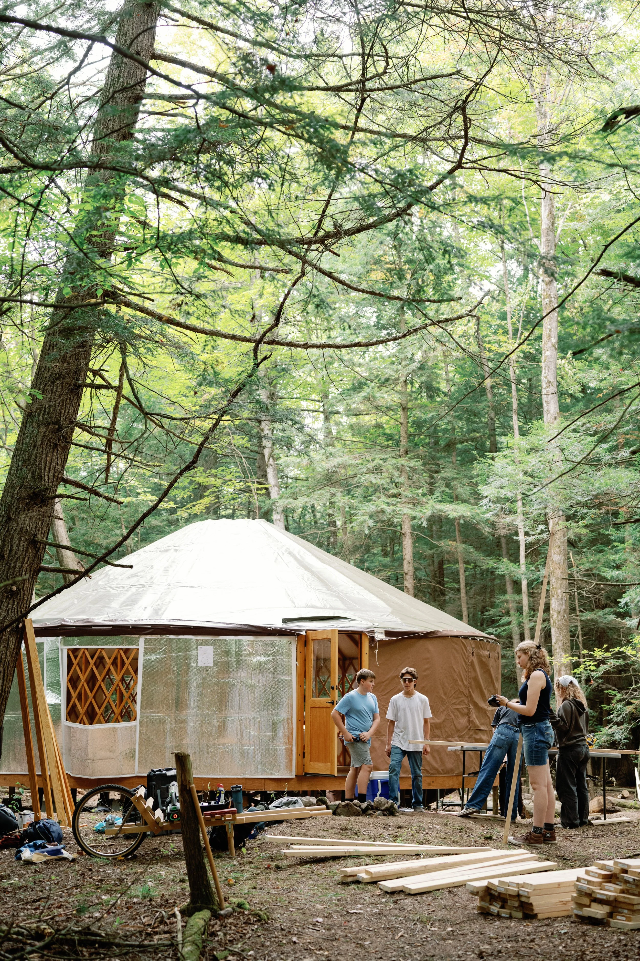 Group of people working on constructing or setting up a large canvas or fabric tent in a forested area, surrounded by trees and nature, with wood planks and tools nearby.