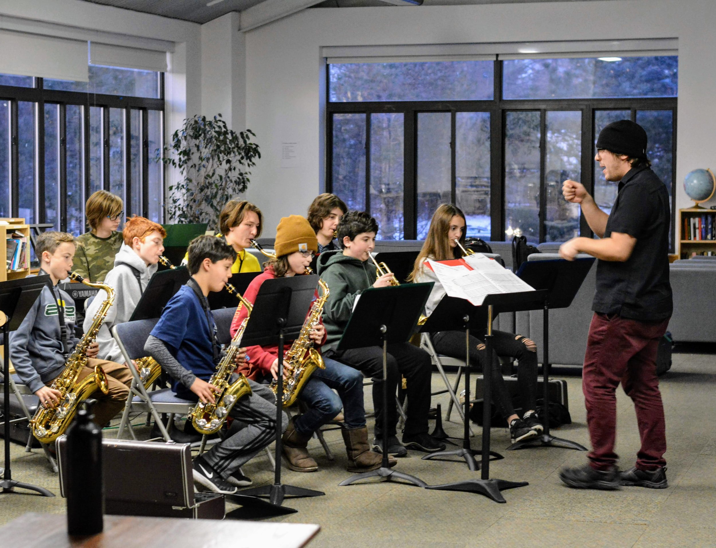 A music instructor leads a group of young students playing saxophones during a band practice in a classroom with large windows and a bookshelf.