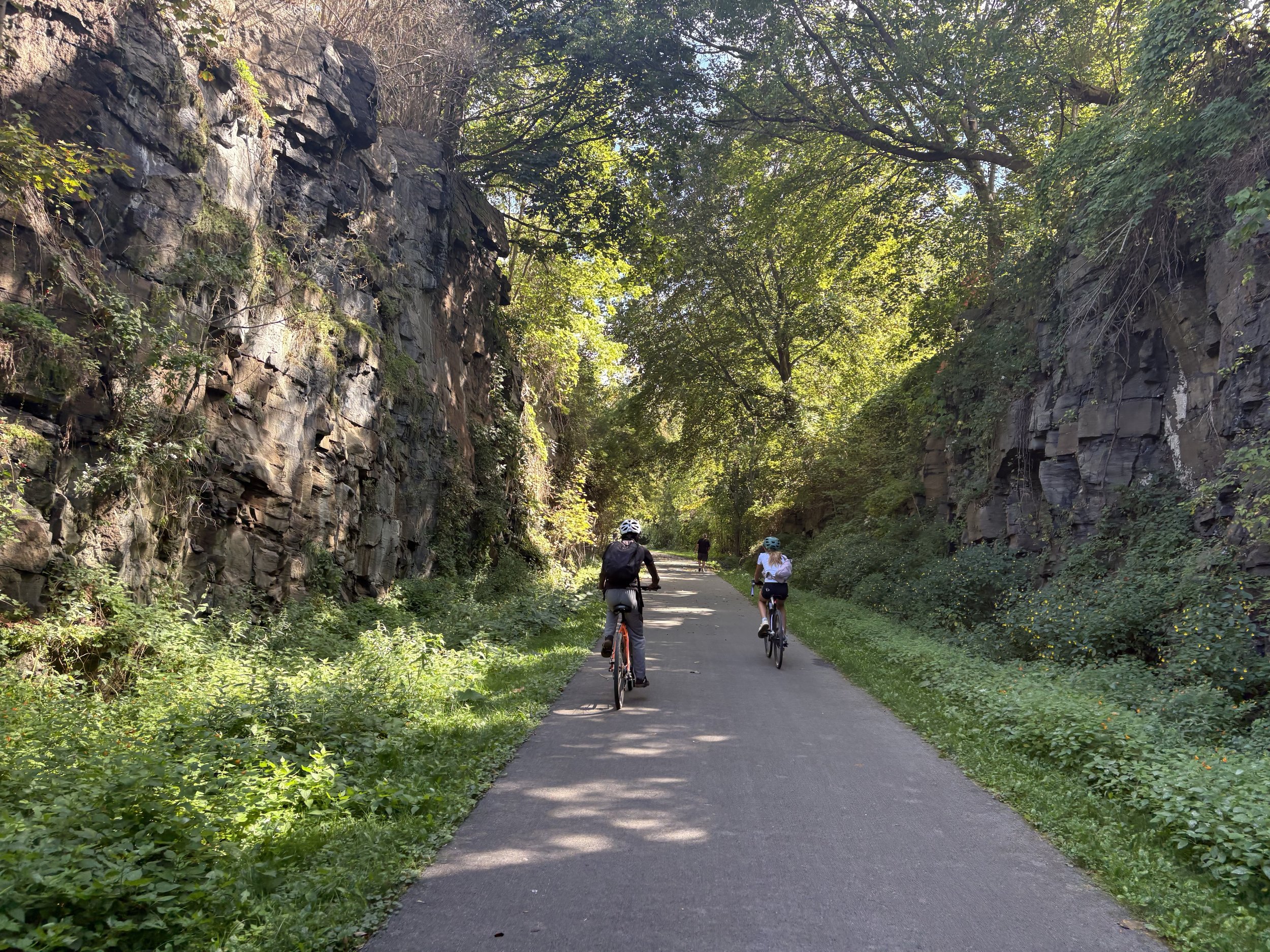 Two cyclists riding on a paved trail through a lush green canyon with rocks and trees overhead.