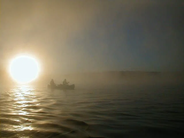 A boat with two people on calm water during sunset or sunrise, with the sun low on the horizon and foggy or misty atmosphere.