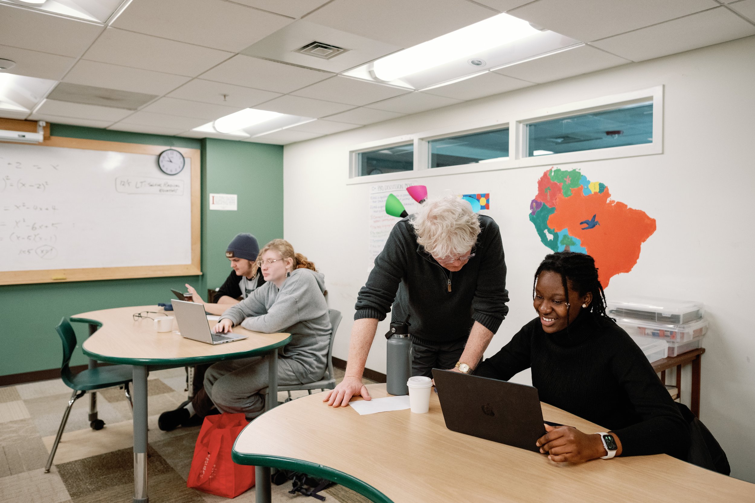 A classroom with four students, one teacher, and colorful South America map on the wall. Three students are working on laptops, one girl and a boy in a hoodie are focused on their screens, and the teacher with curly white hair is assisting a smiling girl in black, who is also using a laptop.