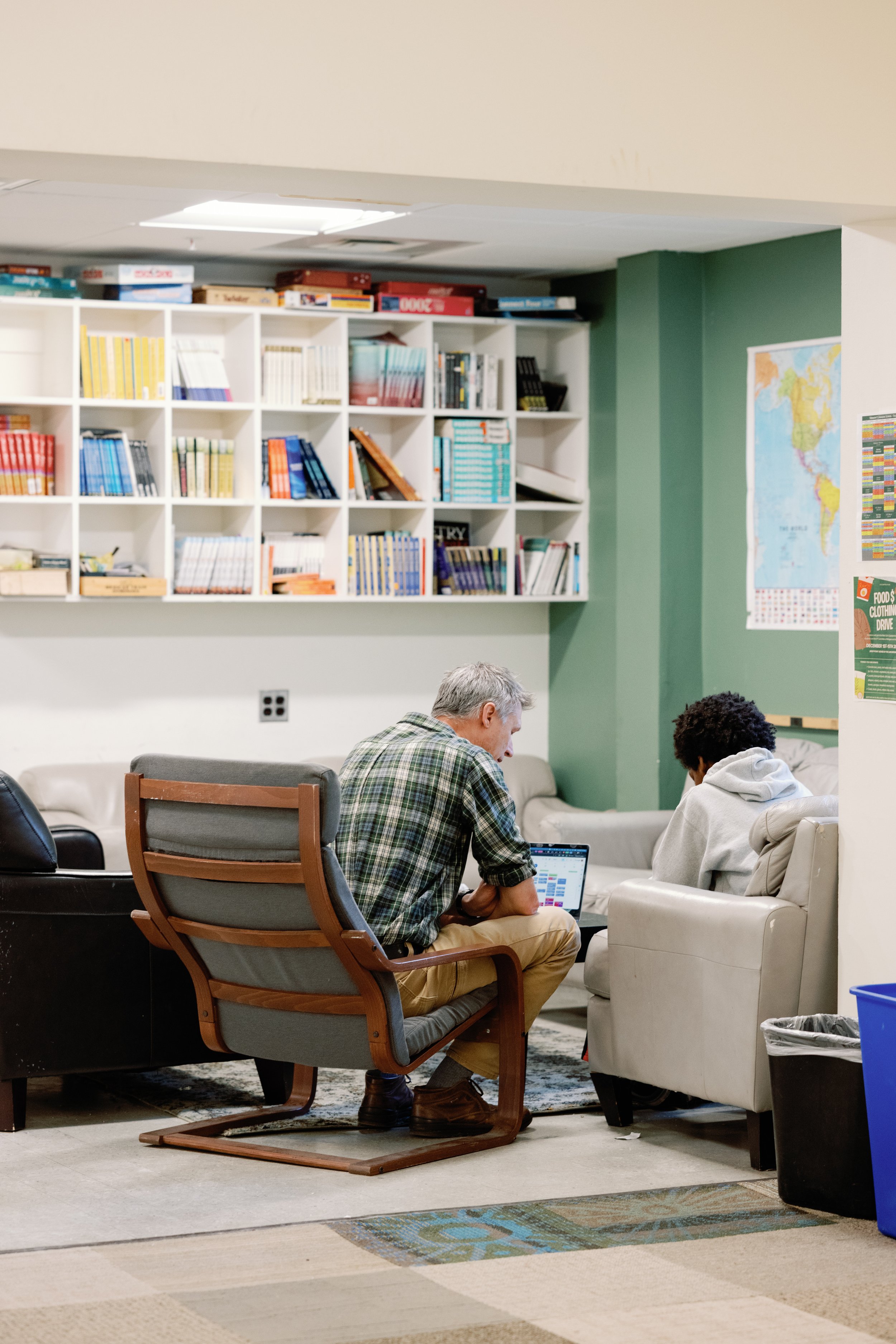 Two people sitting in a cozy room filled with books on white shelves, with a green wall and a world map.