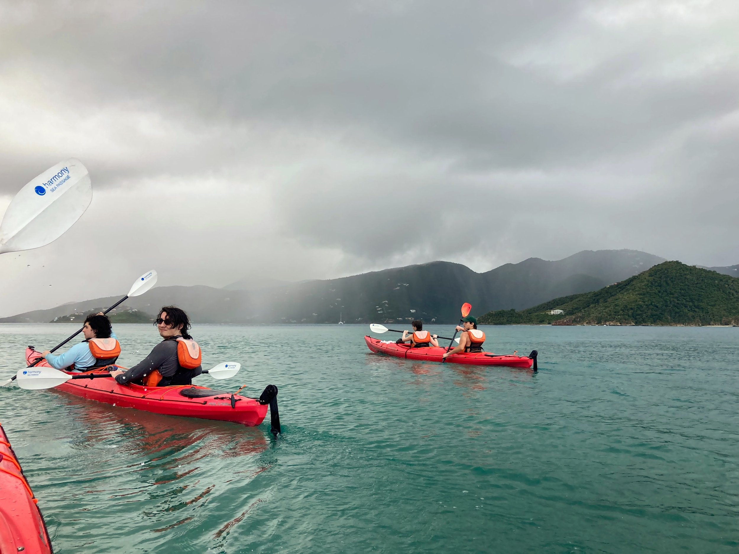 Four people kayaking on a large body of water with hills and overcast sky in the background.