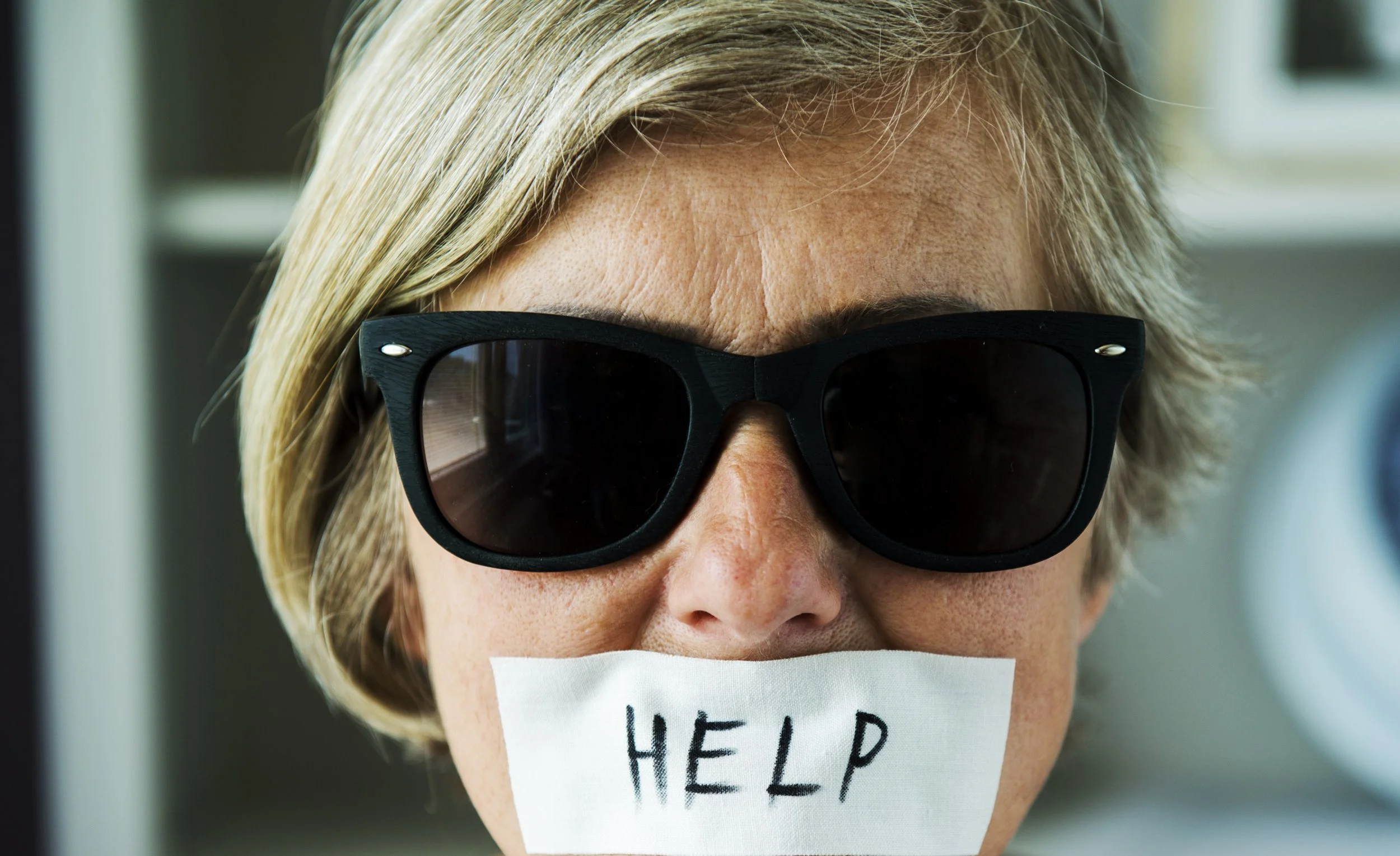 Close-up of a woman with blonde hair wearing large black sunglasses, holding a piece of paper with the word 'HELP' written on it over her mouth.