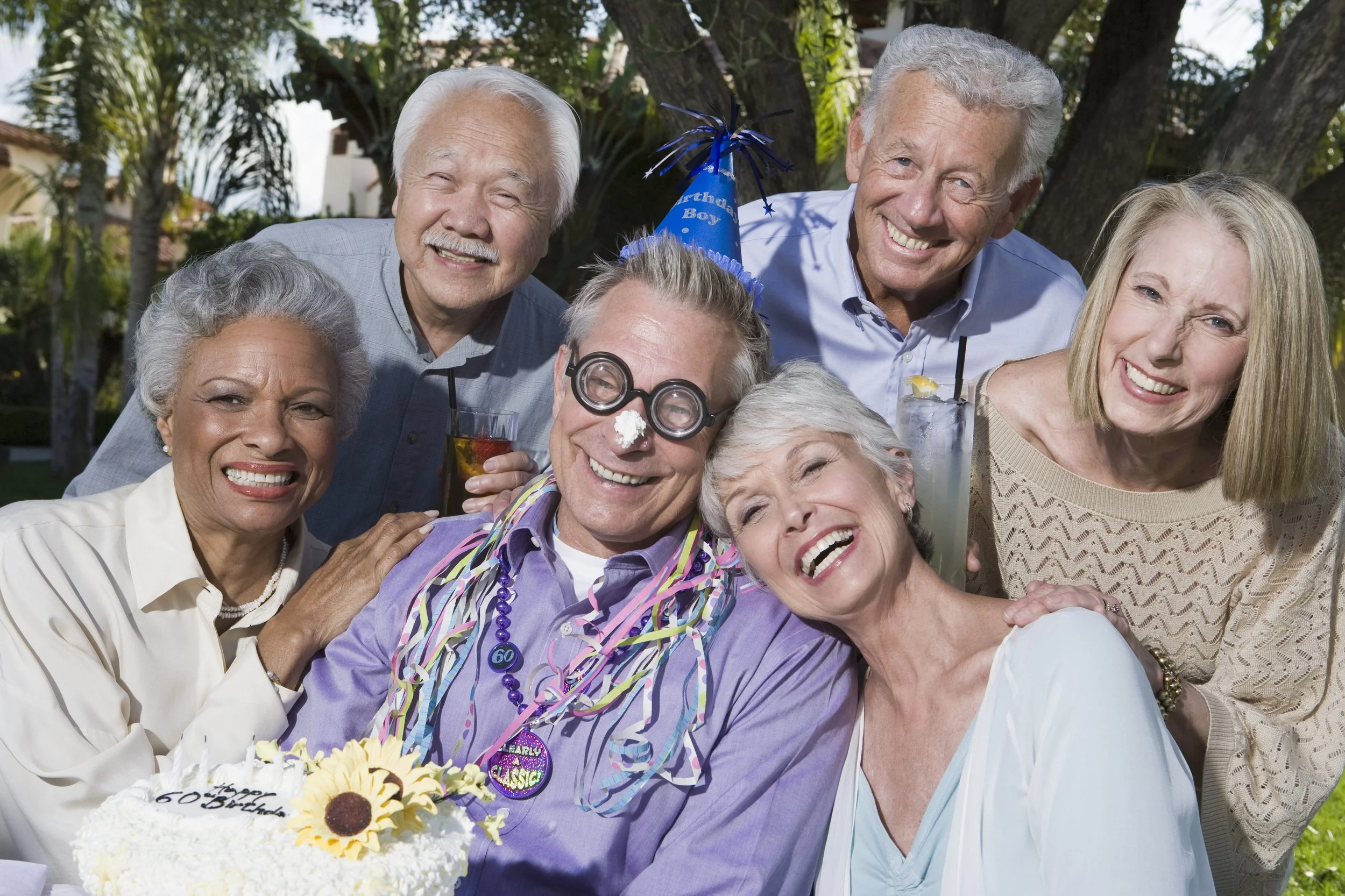 A group of elderly people celebrating a birthday outdoors, with a cake and party hats, all smiling and enjoying the gathering.