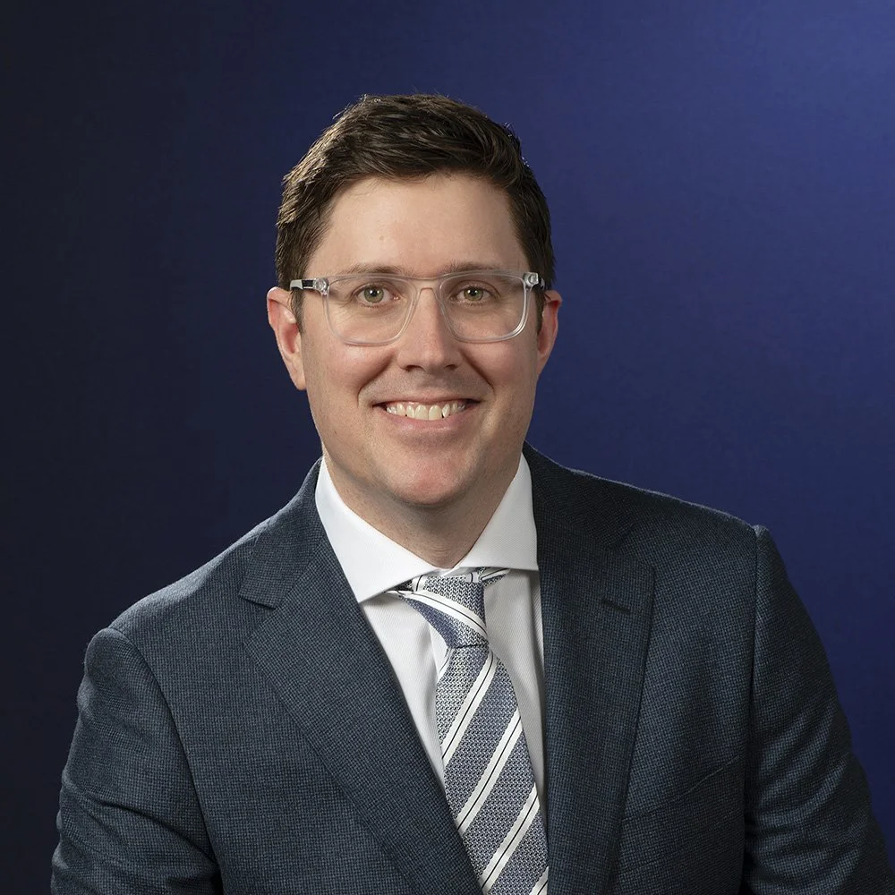 Professional headshot of a man in a dark suit, striped tie, and glasses, smiling against a dark blue background.